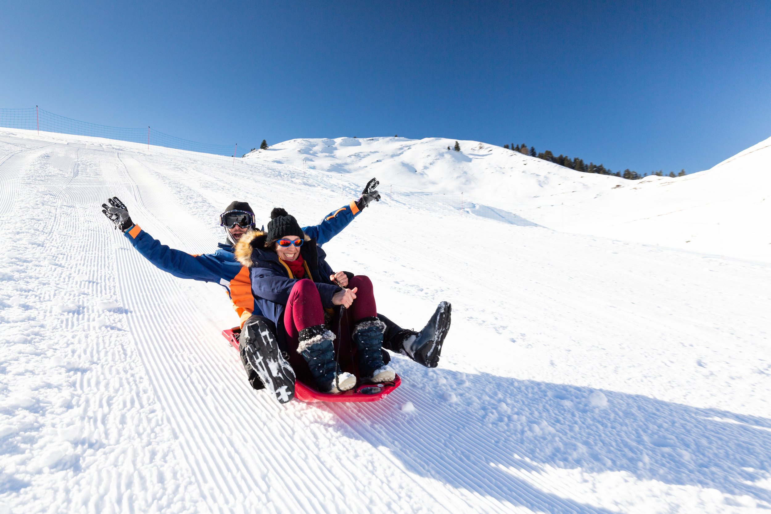 Deux personnes à ski descendent une pente enneigée : une femme et un homme assis sur une luge rouge, souriantes et riant, portant des vêtements d'hiver colorés et un casque de ski, sous un ciel bleu clair.
