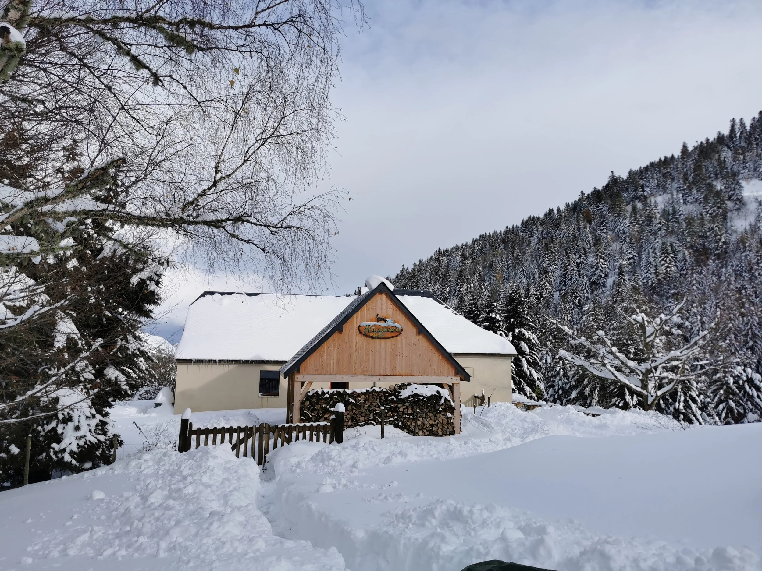 Maison de montagne avec toit enneigé, entourée d'arbres et de montagnes neigeuses, sous un ciel partiellement nuageux.