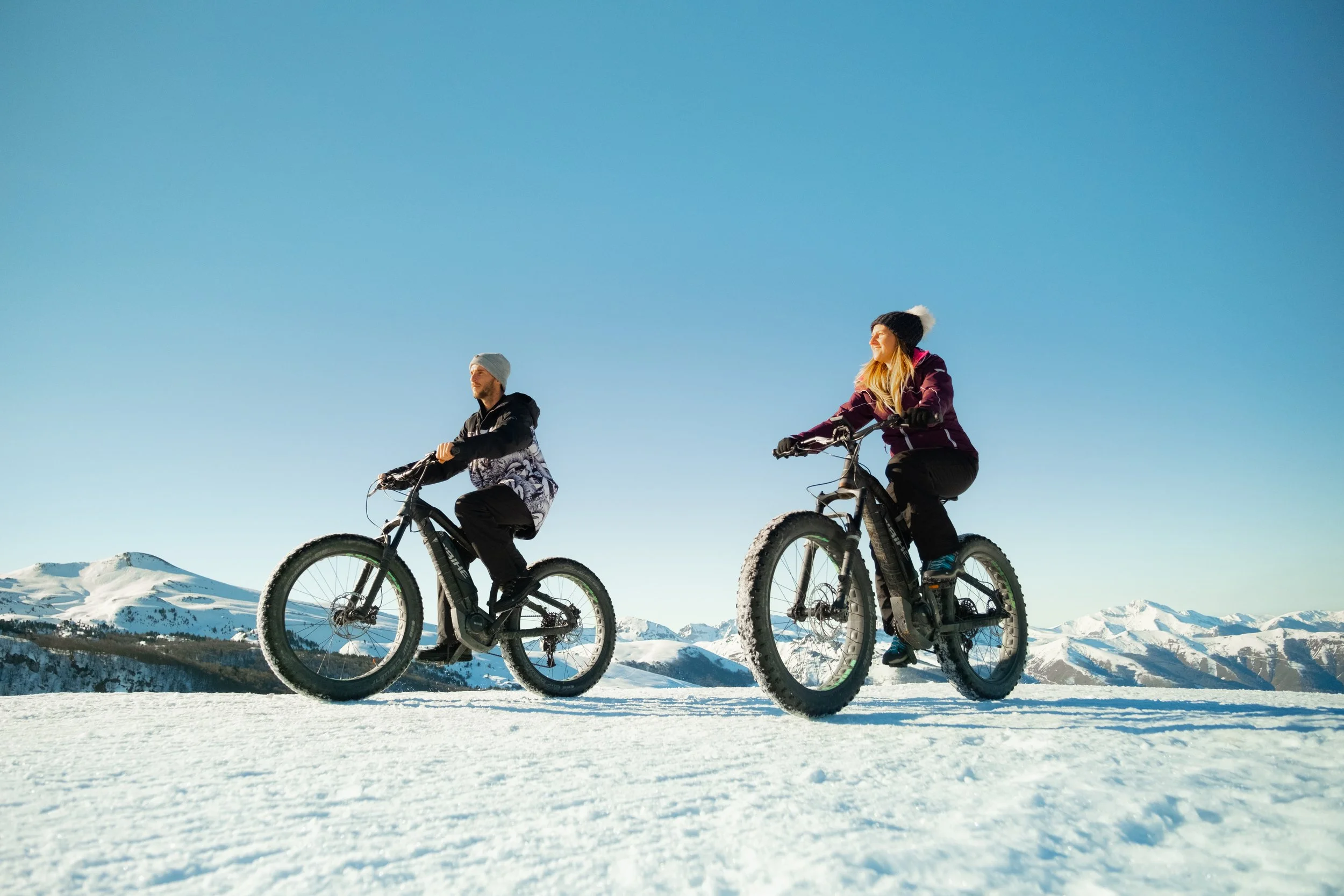 Deux personnes font du vélo dans un paysage enneigé avec des montagnes en arrière-plan, un ciel bleu clair.