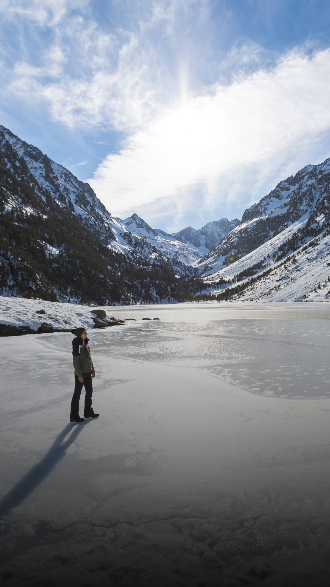 Une personne debout sur un lac gelé entouré de montagnes enneigées sous un ciel partiellement nuageux.