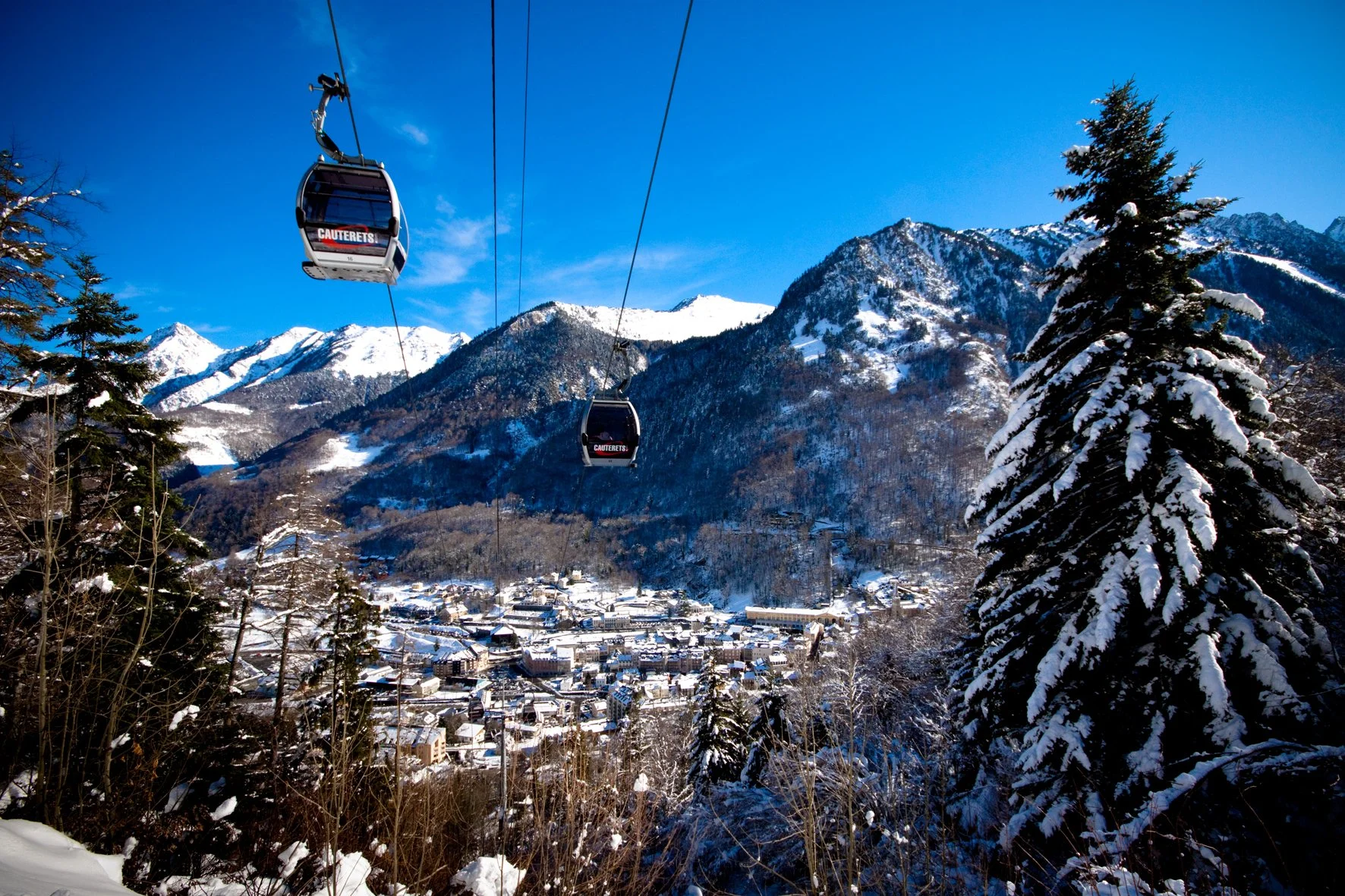 Deux téléphériques survolent une station de ski enneigée dans un paysage de montagnes avec un ciel bleu clair.