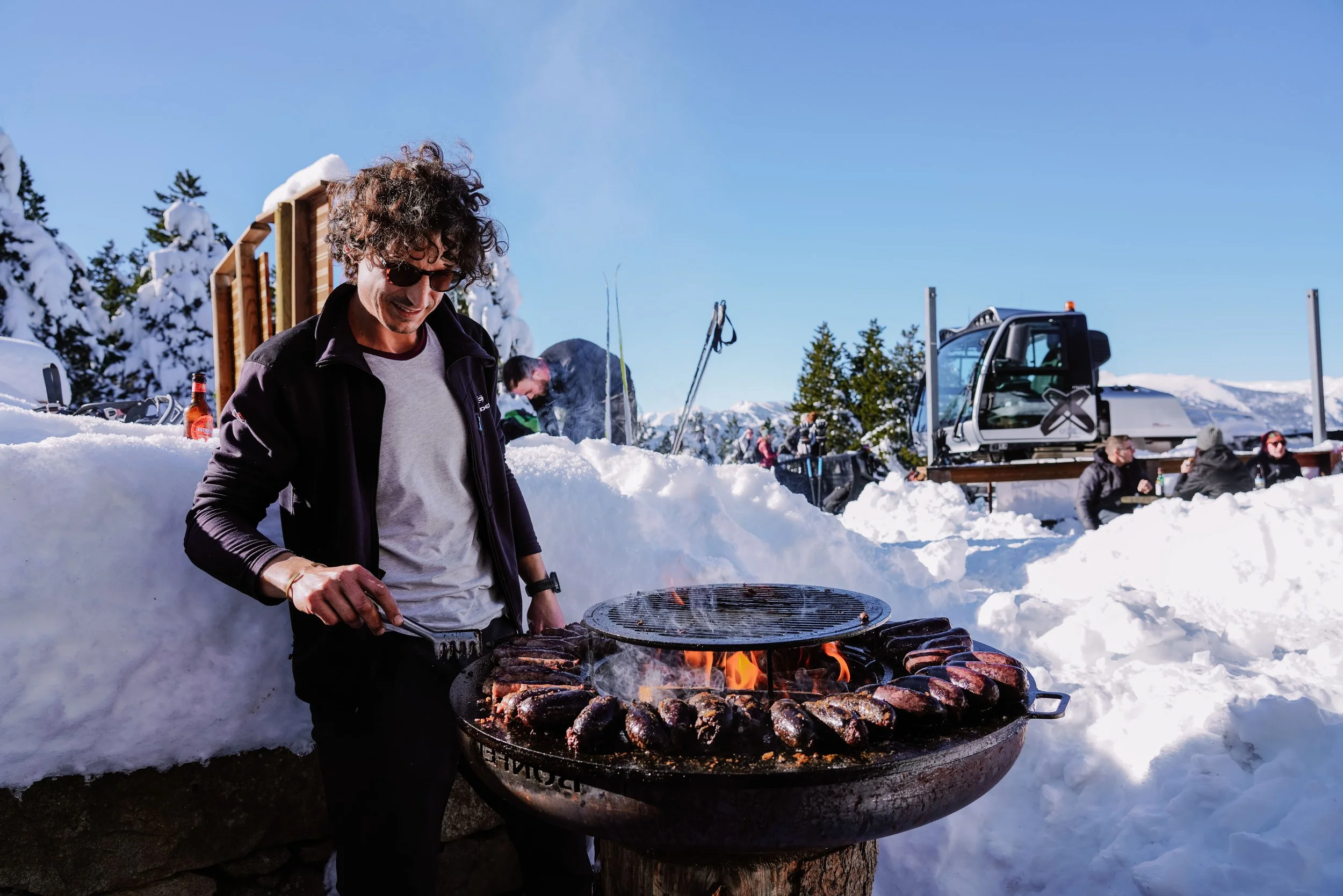 Une personne fait griller des viandes sur un barbecue en plein air dans un paysage enneigé, avec d'autres personnes autour, en arrière-plan, en montagne et sous un ciel clair.