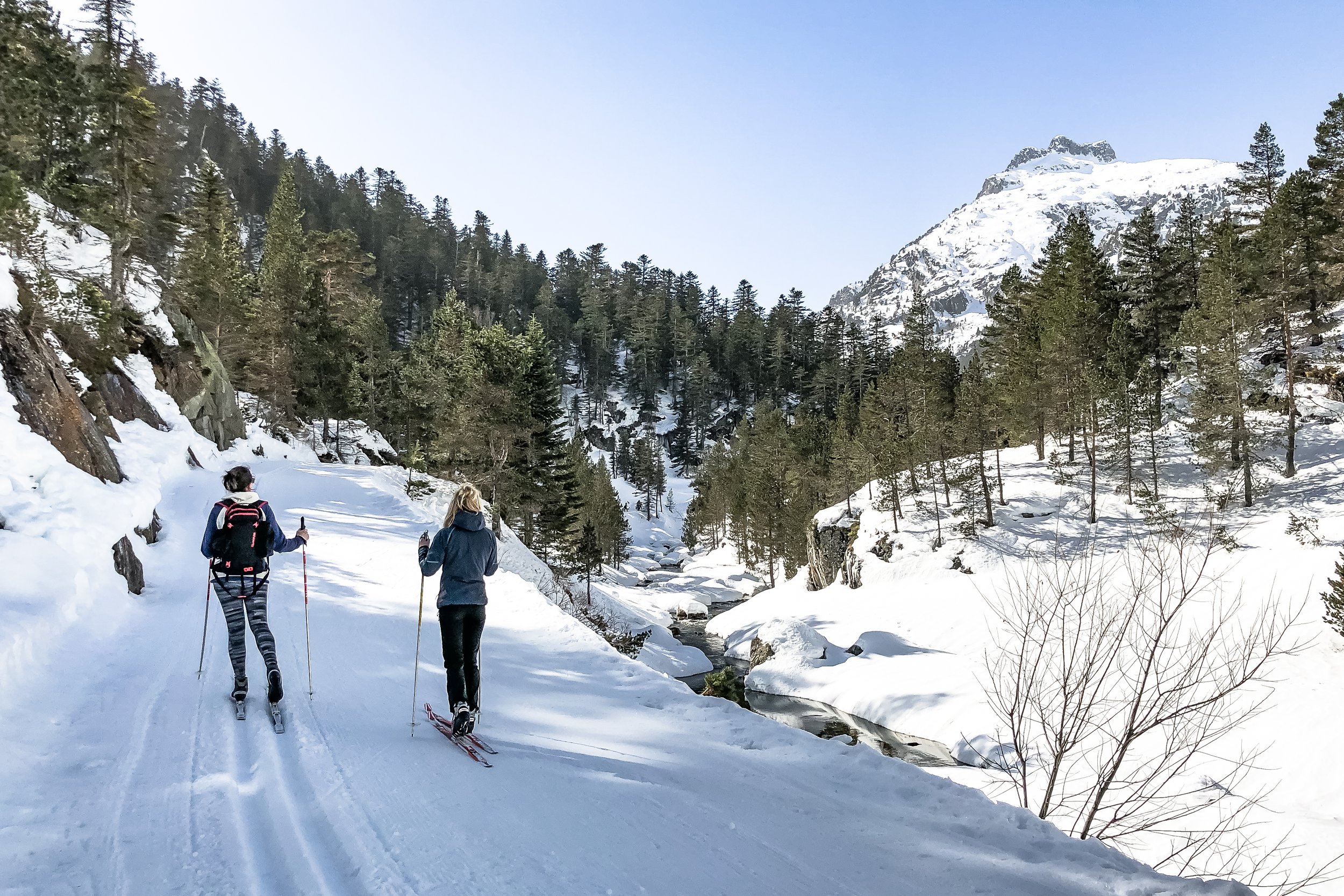 Deux personnes faisant du ski de fond dans une vallée enneigée entourée de pins et de montagnes, par une journée ensoleillée.