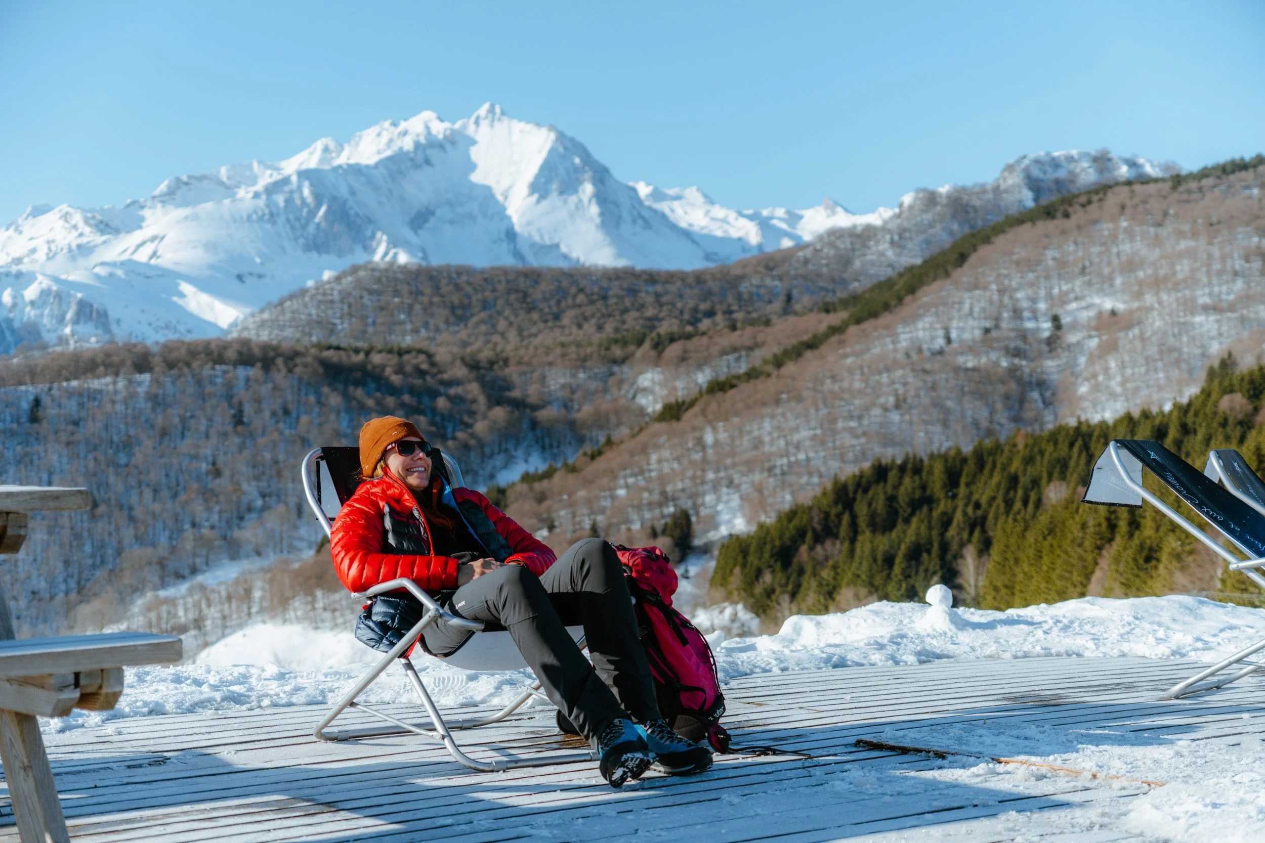 Une personne souriante repose dans une chaise pliante sur une terrasse enneigée, avec des montagnes enneigées en arrière-plan ensoleillé.