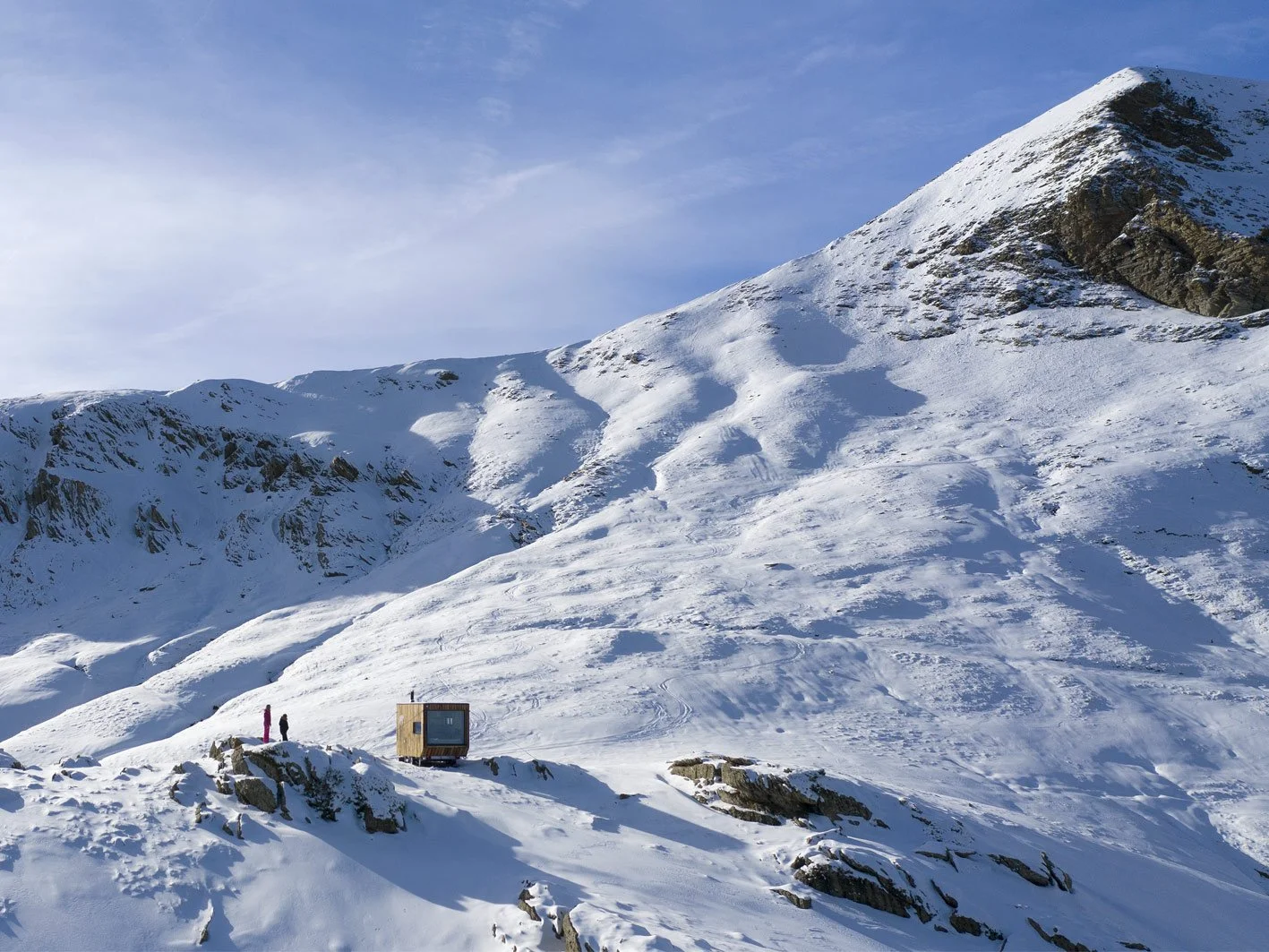 Montagne enneigée avec deux personnes et une petite cabane en bois près du bas, ciel partiellement nuageux.