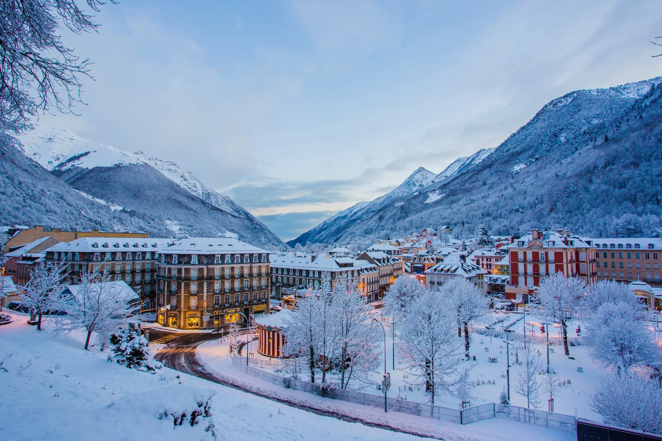 Ville enneigée dans une vallée montagneuse, avec bâtiments, arbres et rues recouverts de neige, sous un ciel nuageux.