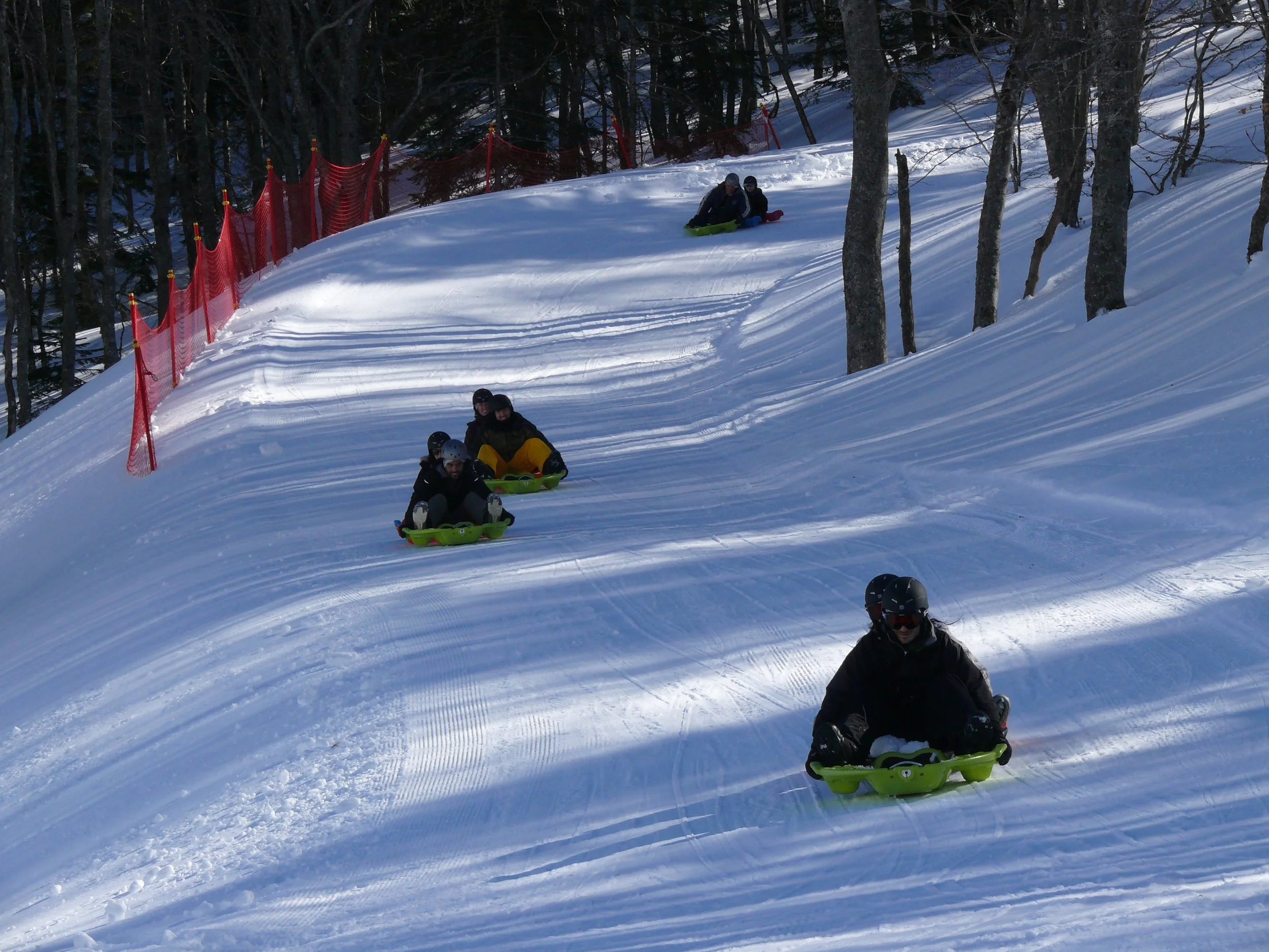 Groupe de personnes faisant de la luge sur une pente enneigée bordée d'arbres et de filets de protection rouges.