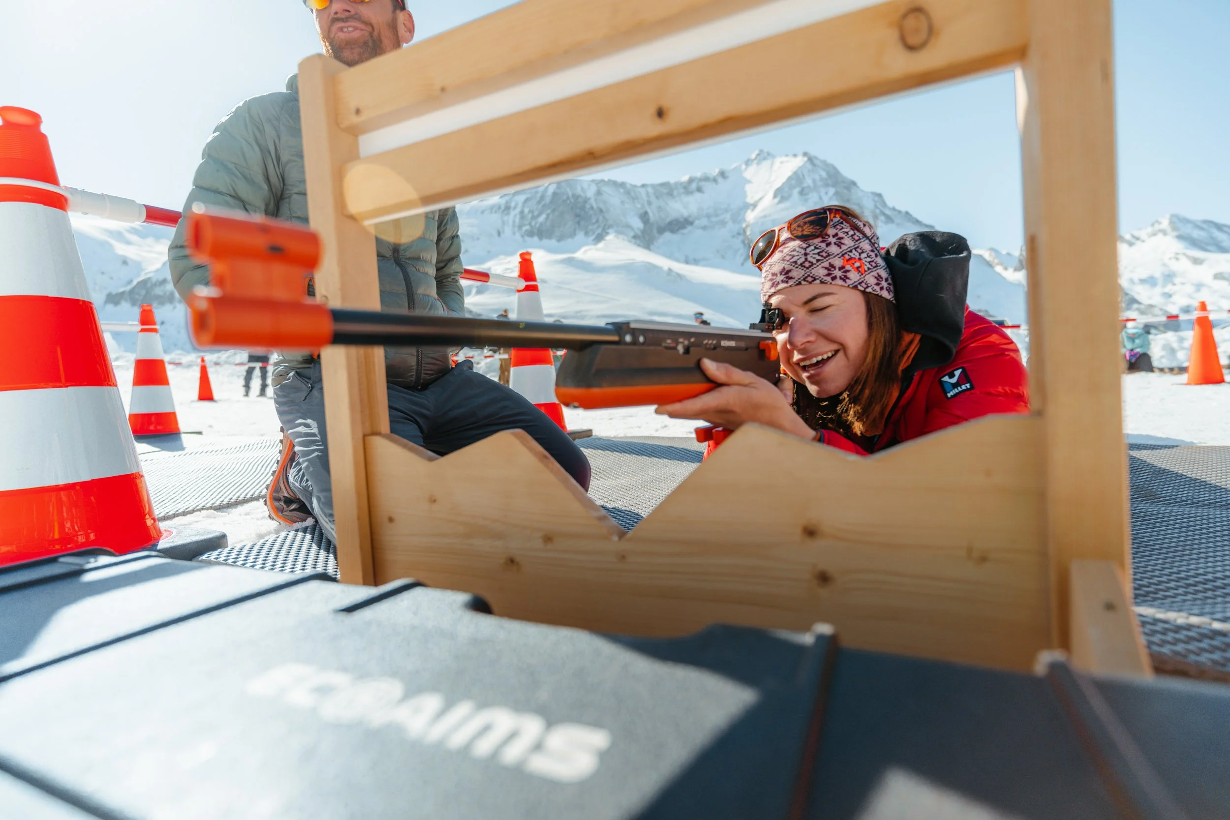 Une femme souriante, en ski, regarde dans une lunette de tir à air comprimé dans un environnement enneigé avec des montagnes en arrière-plan.