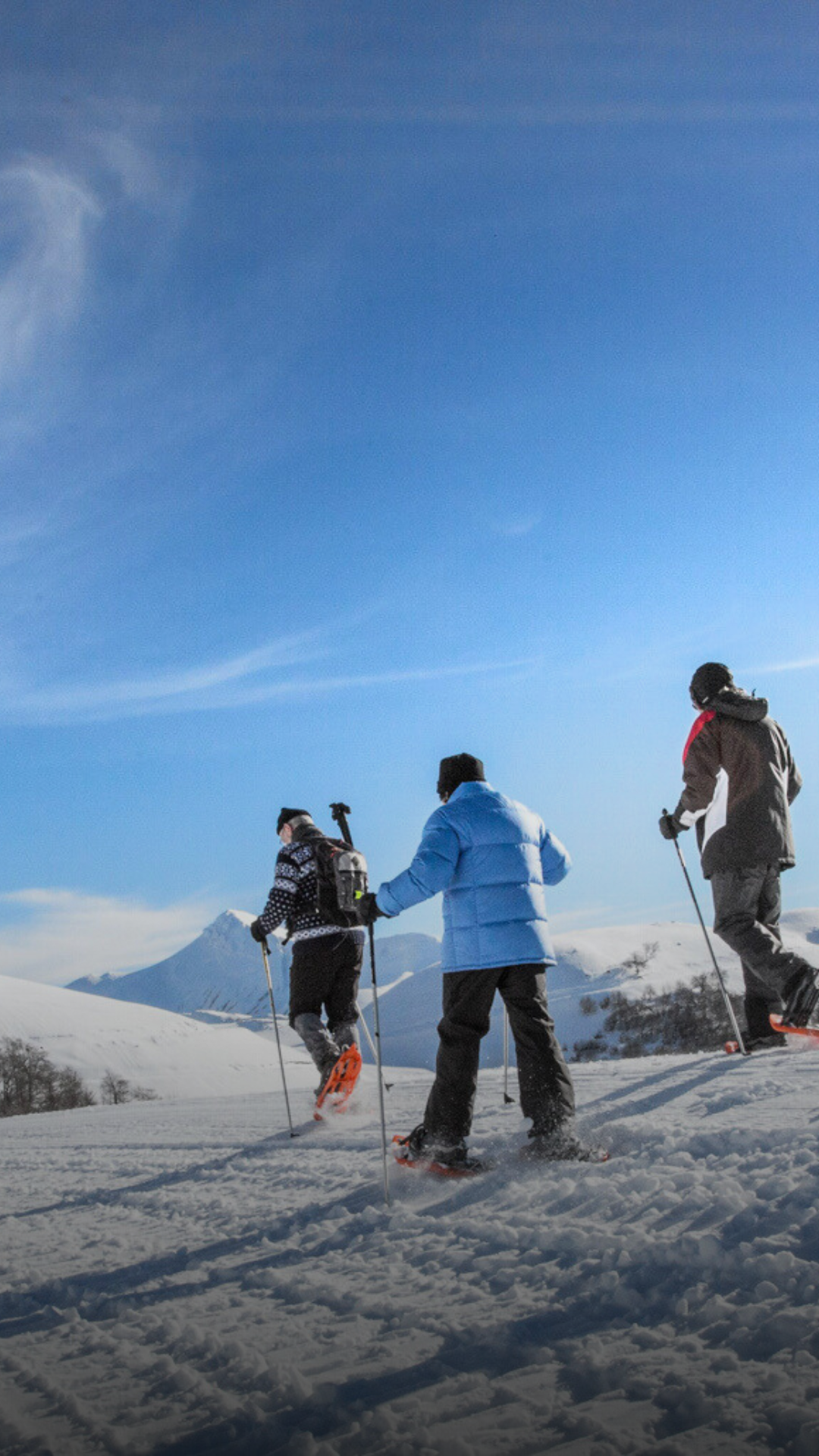 Trois personnes en ski dans un paysage enneigé avec des montagnes en arrière-plan et un ciel bleu clair.