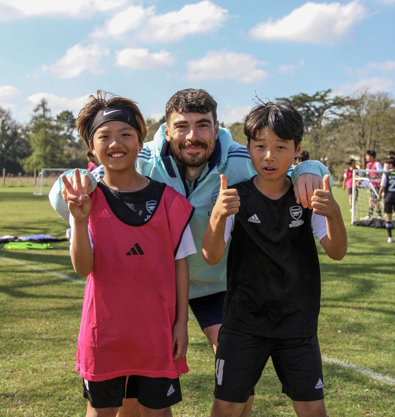 サッカーの練習をしている少年二人と男性監督の集合写真。少年たちは勝利のポーズとサムズアップをしている。青空と緑のフィールドが背景。