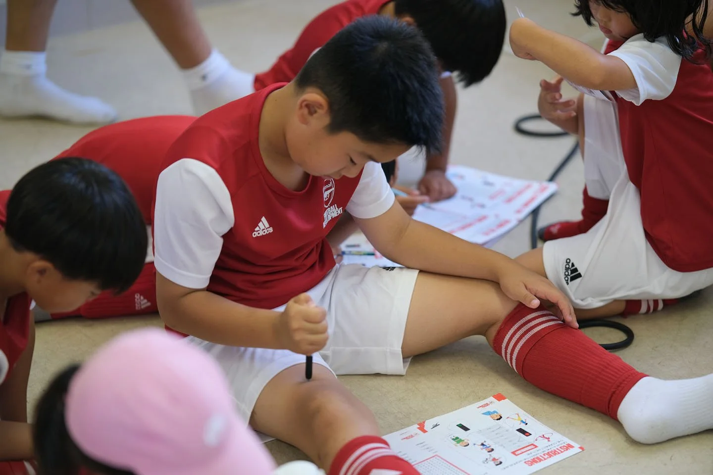 体育の授業を行っている子どもたちと先生。子供たちは赤いユニフォームを着て、地面に座っている。