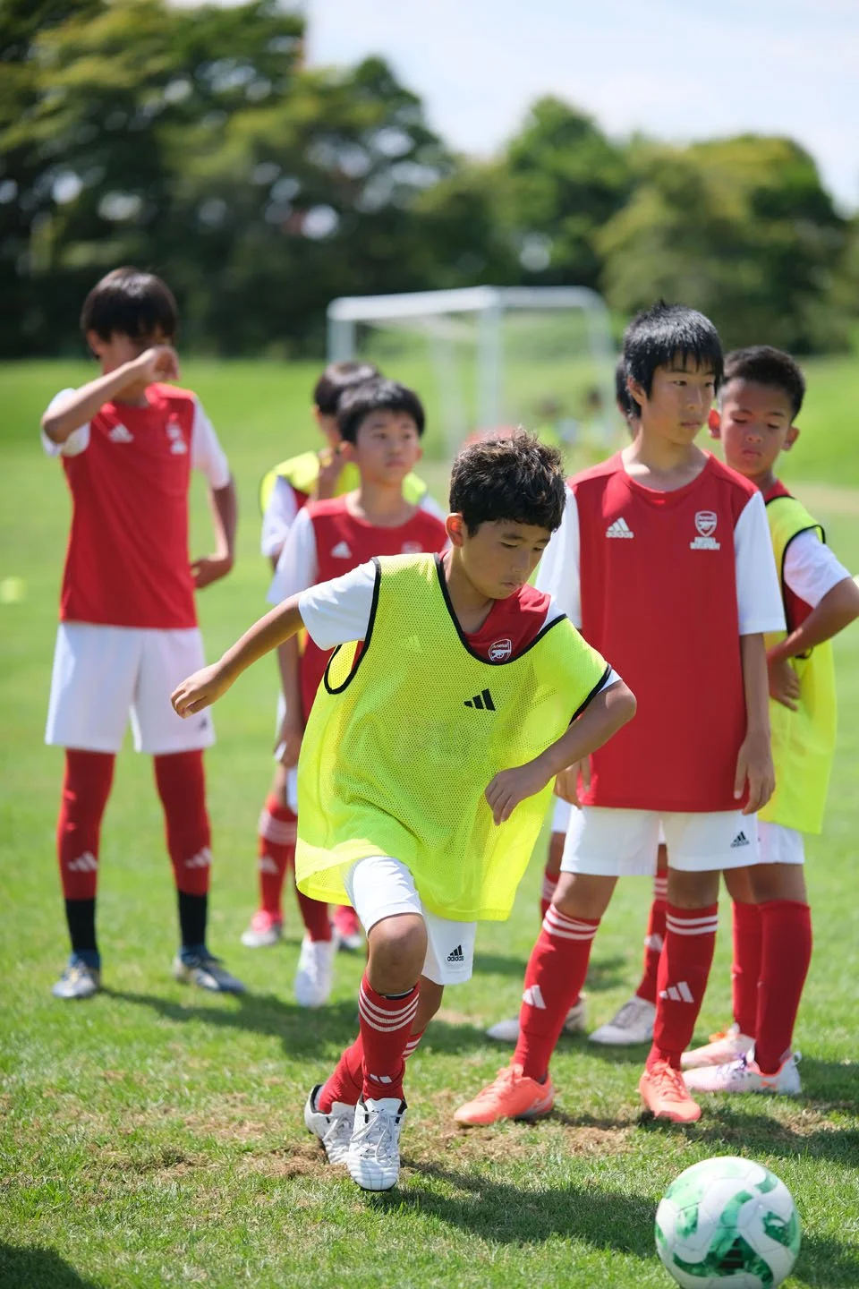 サッカーチームの子供たちが練習している。少年が緑のゴールキックの練習をしているところ。背景に木々と青空が見える。