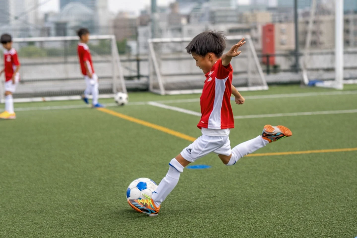 サッカーの試合をしている子供たち。少年が赤いユニフォームを着て、緑のサッカー場でボールを蹴っている。背景にはゴールと他の子供たちがいる。