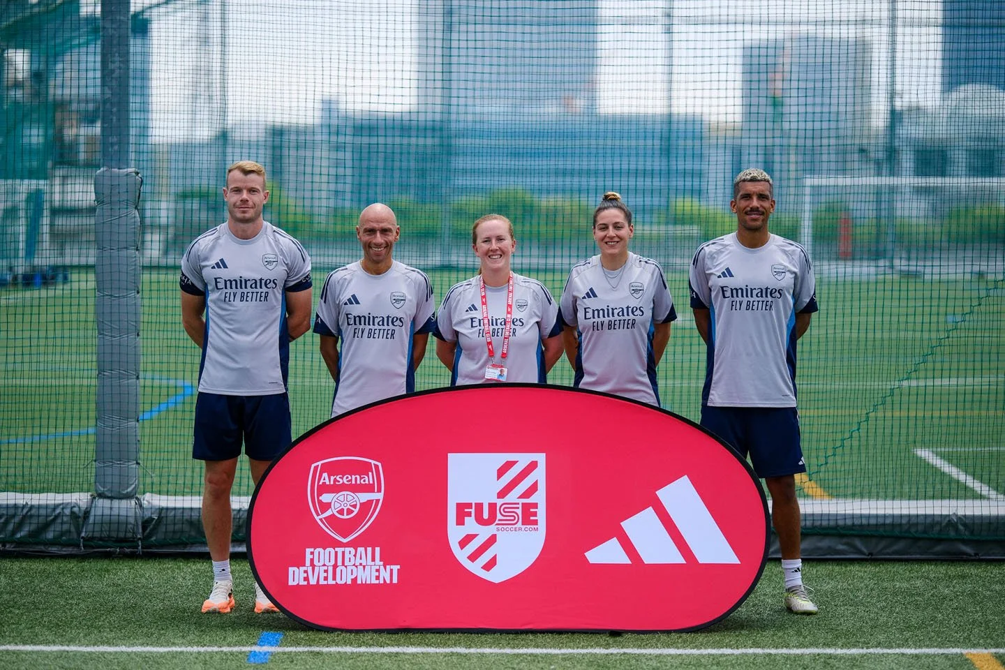 サッカートレーニング中の男女5人のグループ写真。背景はサッカー場のネットと都市の高層ビル。前には「FOOTBALL DEVELOPMENT」の文字とアーセナルのロゴを含む旗が立っている。