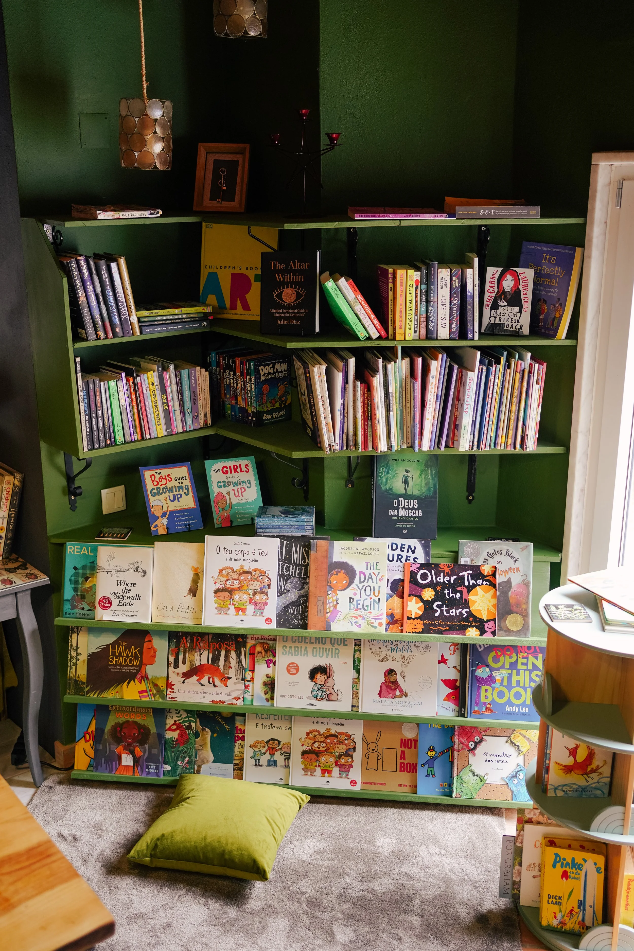 A green bookshelf filled with children's books, with some books arranged upright and others stacked horizontally. On the shelf, there are also a few framed pictures and decorative items. A green pillow is on the floor in front of the bookshelf.