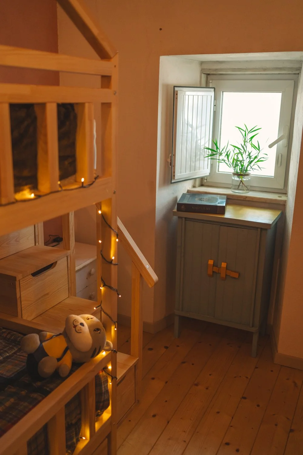A cozy corner of a room with a wooden staircase decorated with string lights, a small stuffed toy on a plaid bedcover, a small cabinet near a window, and a potted plant on the window sill.