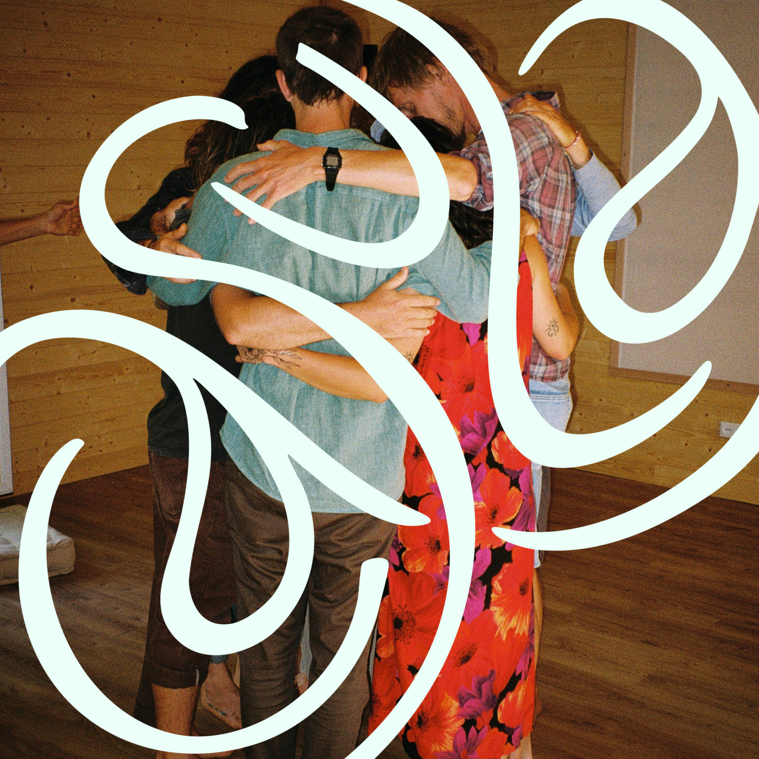 Group of people embracing in a circle indoors, with a wooden wall background and wooden floor, some wearing casual clothes.