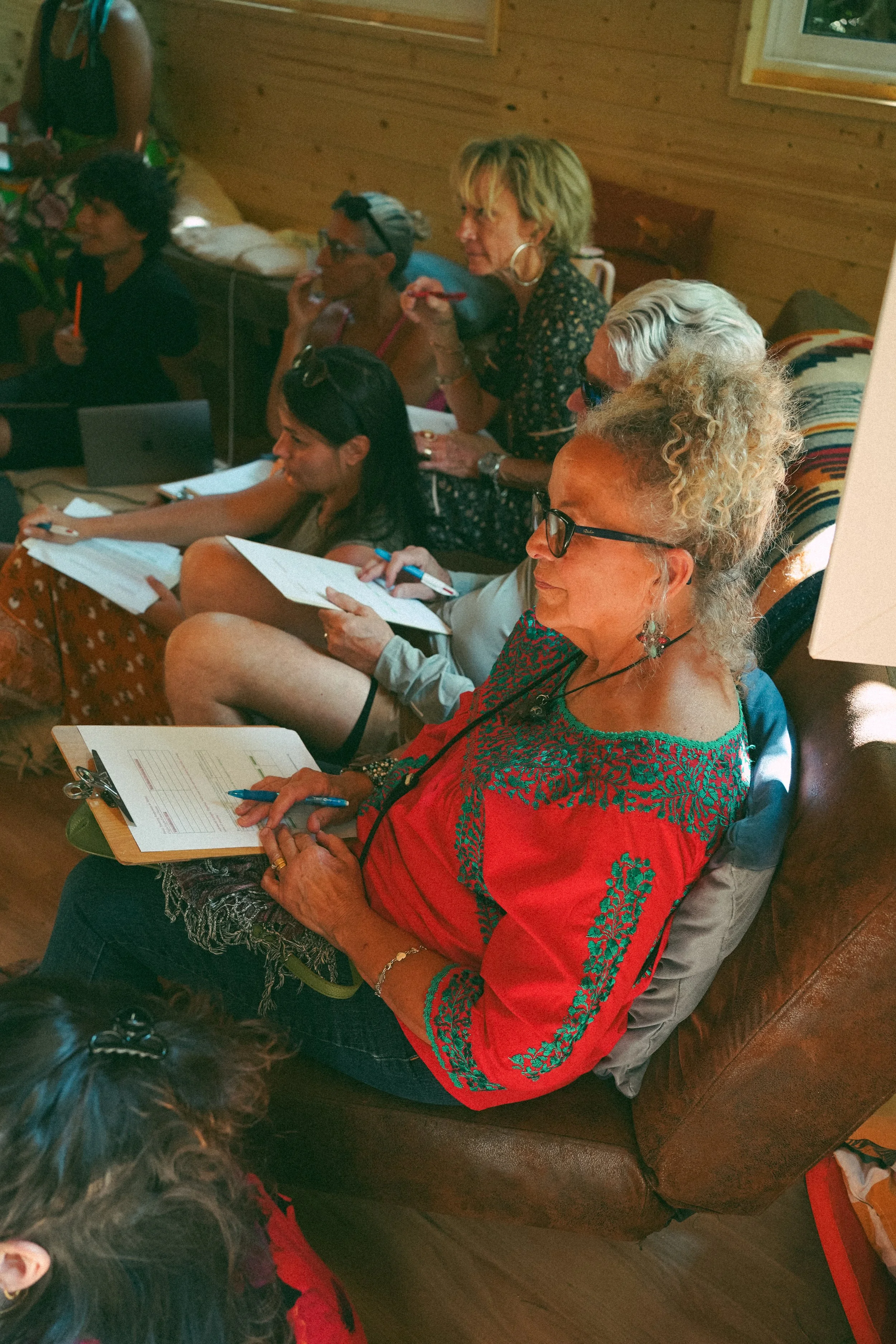 A group of diverse women sitting on a leather couch, taking notes and listening attentively in an indoor setting with wooden walls.
