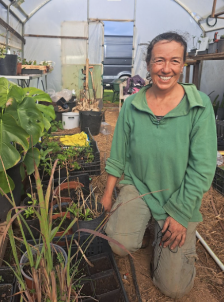 Smiling woman wearing a green hoodie and beige pants kneeling inside a greenhouse surrounded by plants and gardening supplies.