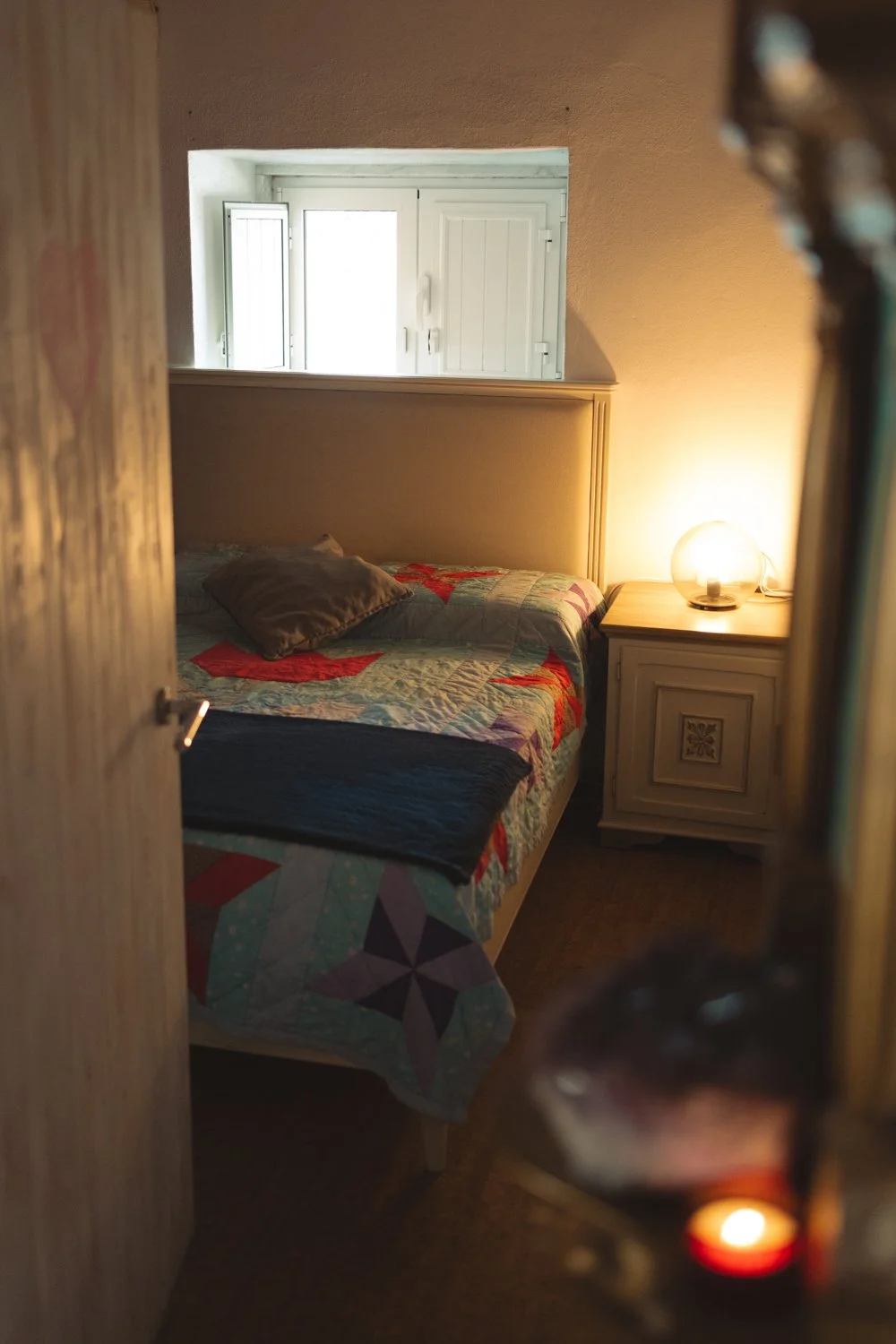 A cozy bedroom viewed through a partially open door, with a bed covered with a colorful quilt, a single pillow, a nightstand with a lit lamp, and a small window with closed shutters.