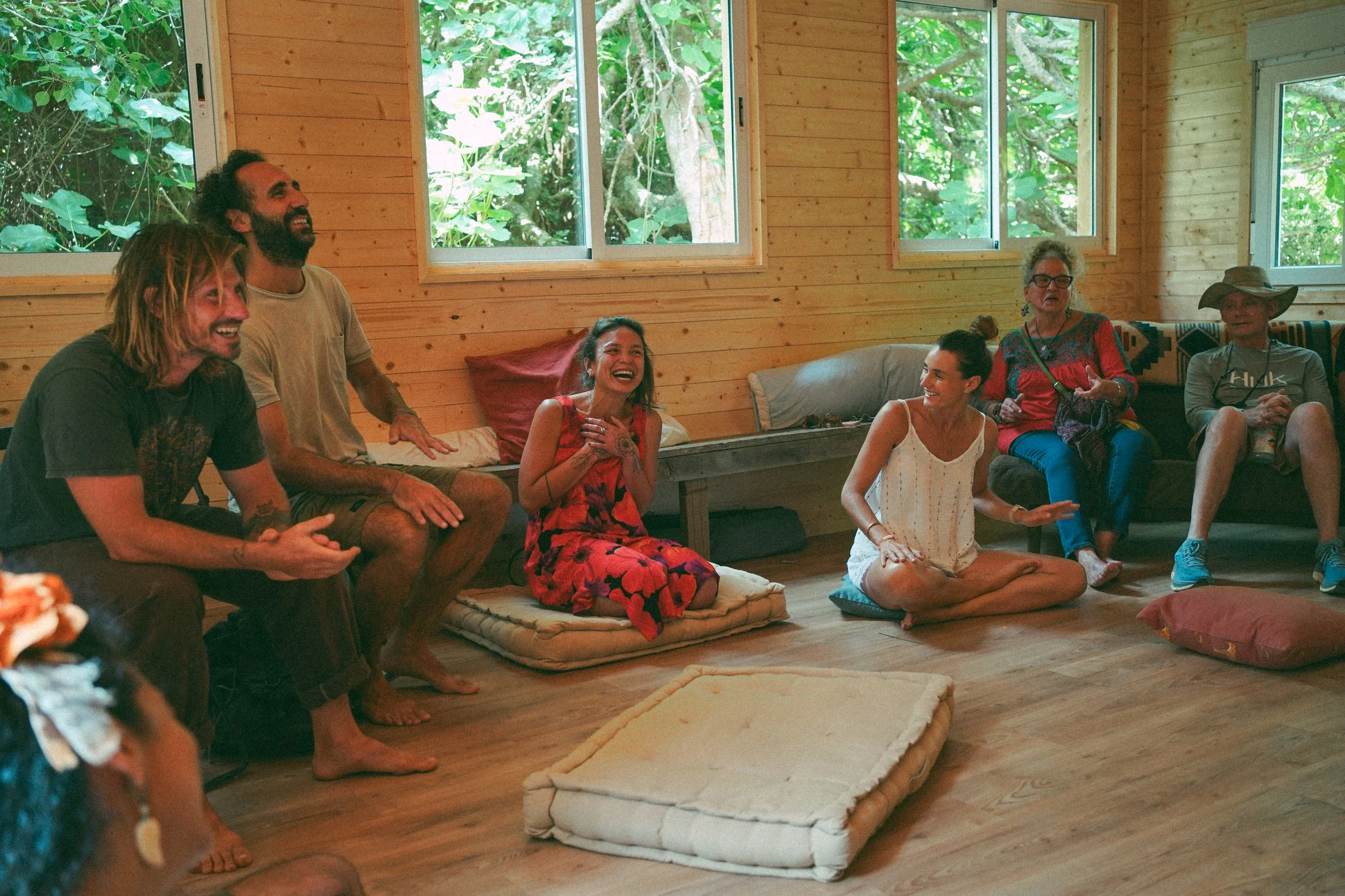 Group of people sitting on cushions in a wooden room with large windows, laughing and enjoying each other's company.
