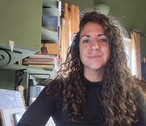 A woman with curly hair and a nose piercing smiling at the camera in a room with green walls, shelves with books, and an orange curtain.