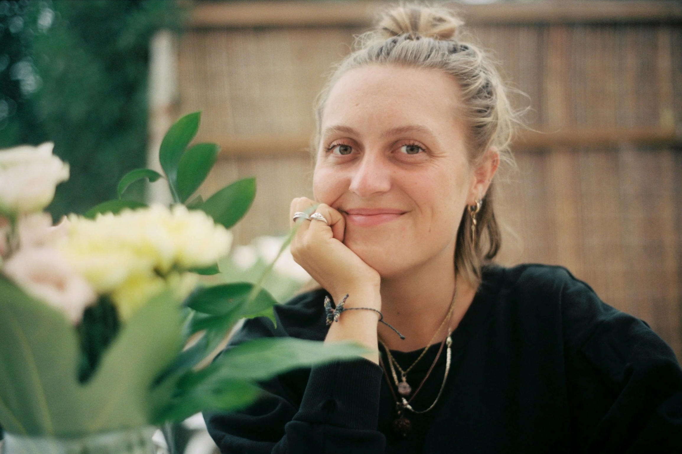 A woman with blonde hair and hoop earrings, smiling while resting her chin on her hand, sitting near a vase of yellow and white flowers, in a cozy indoor setting.