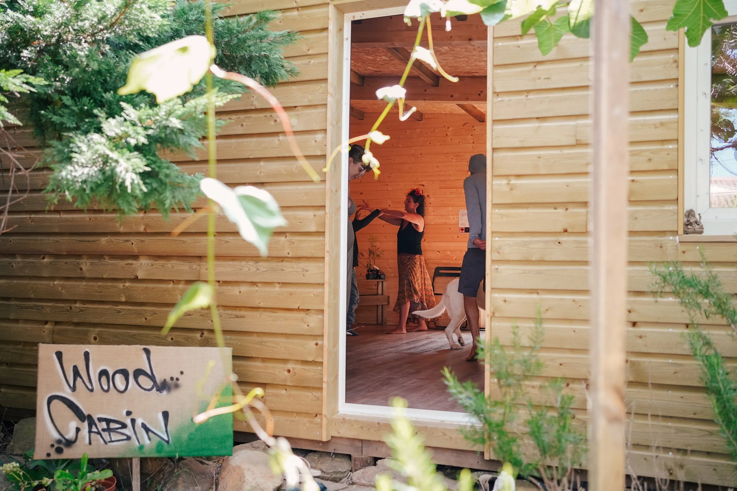 View of a wooden tiny house with an open door, showing people inside celebrating, with a woman dancing and someone holding a dog on a leash