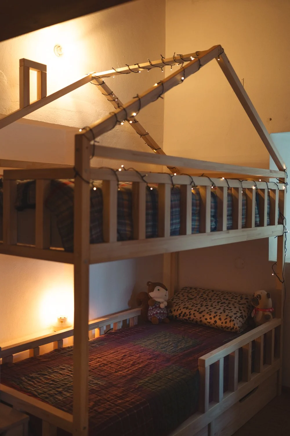 A wooden bed with a loft structure, decorated with string lights. The lower bed has a plaid quilt, a leopard print pillow, and stuffed toys, while the upper bed has a plaid cushion and appears to have a pillow or blanket. The room is warmly lit with wall-mounted and table lamps.