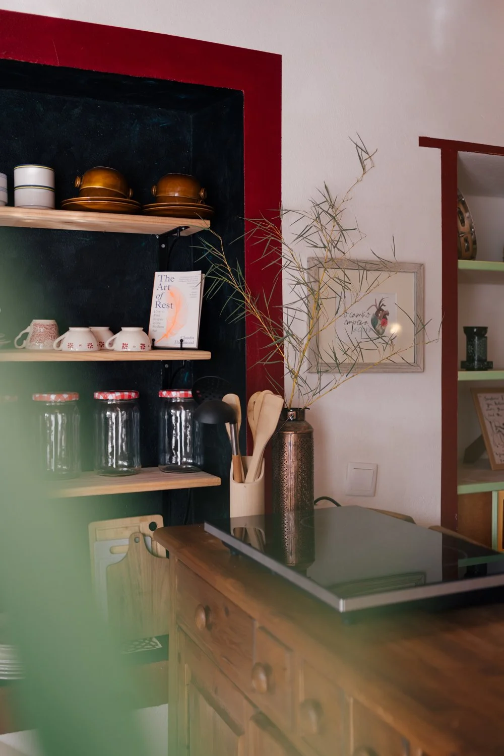 A cozy kitchen corner with wooden shelves holding jars, cups, and teapots. A framed botanical print hangs on the wall, and a tall vase with branches sits on a wooden countertop.