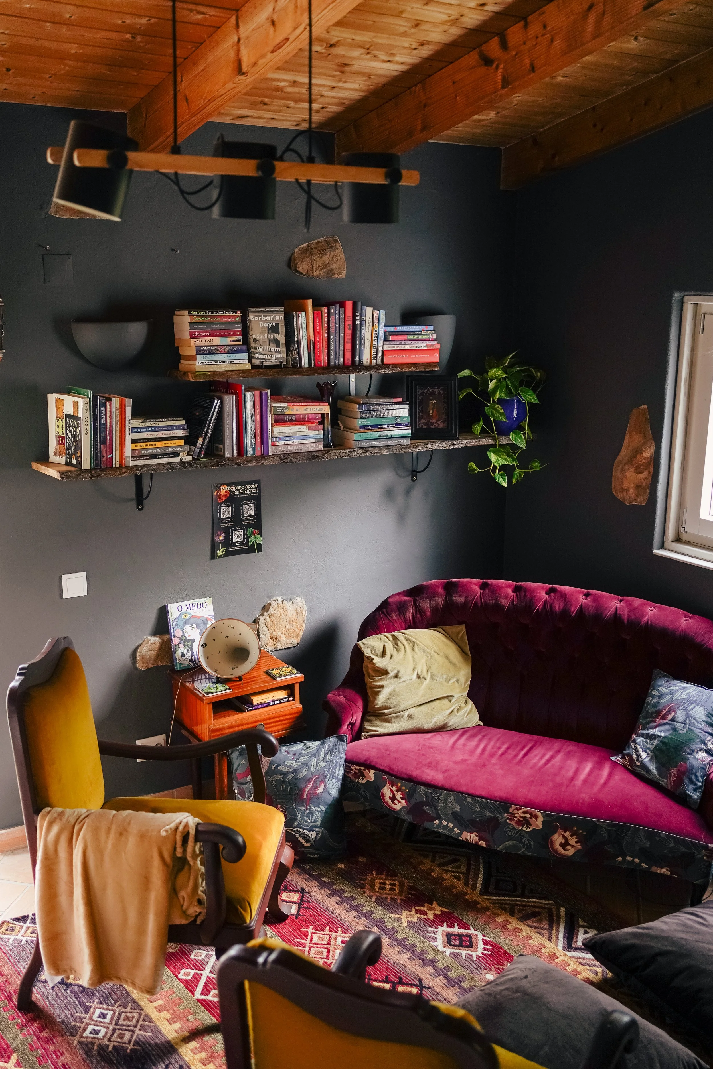 A cozy living room with dark gray walls, wooden ceiling beams, a pink velvet sofa with patterned pillows, a yellow armchair with a blanket, and a wooden shelf filled with books, potted plants, and decorative items. There is a window letting in natural light.