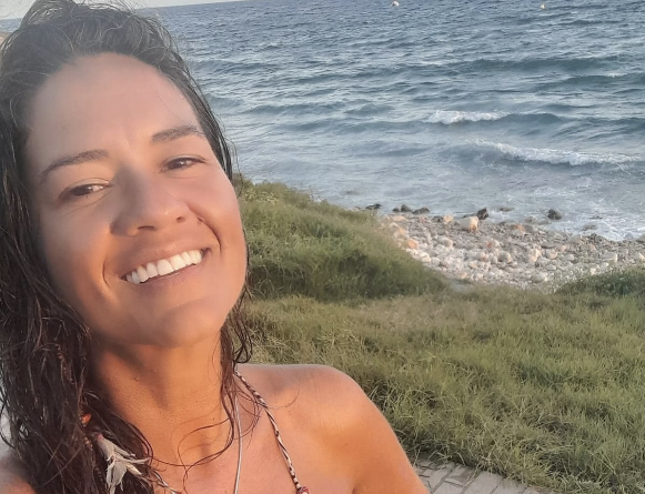 A woman smiling at the camera with the ocean and rocky shoreline in the background.