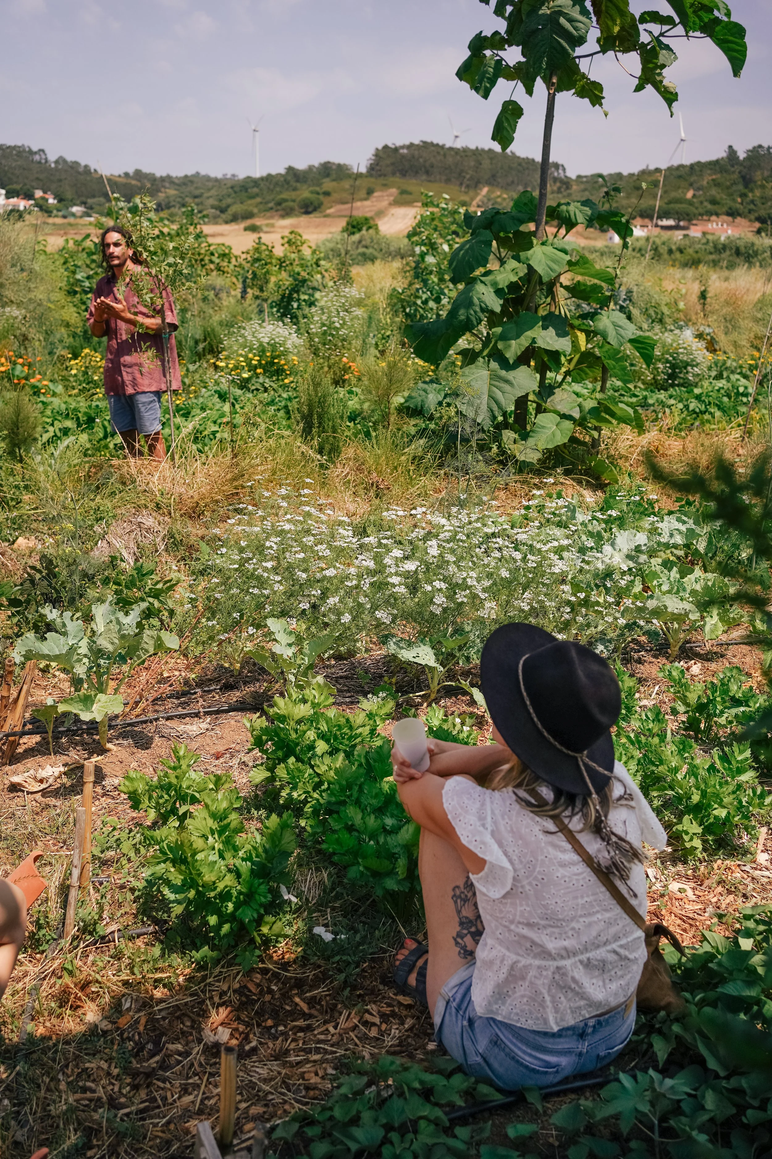 A woman sitting on the ground, wearing a black hat and white blouse, is watering plants in a lush garden or farm. In the background, a man is standing among various plants, with hills, wind turbines, and a partly cloudy sky behind them.