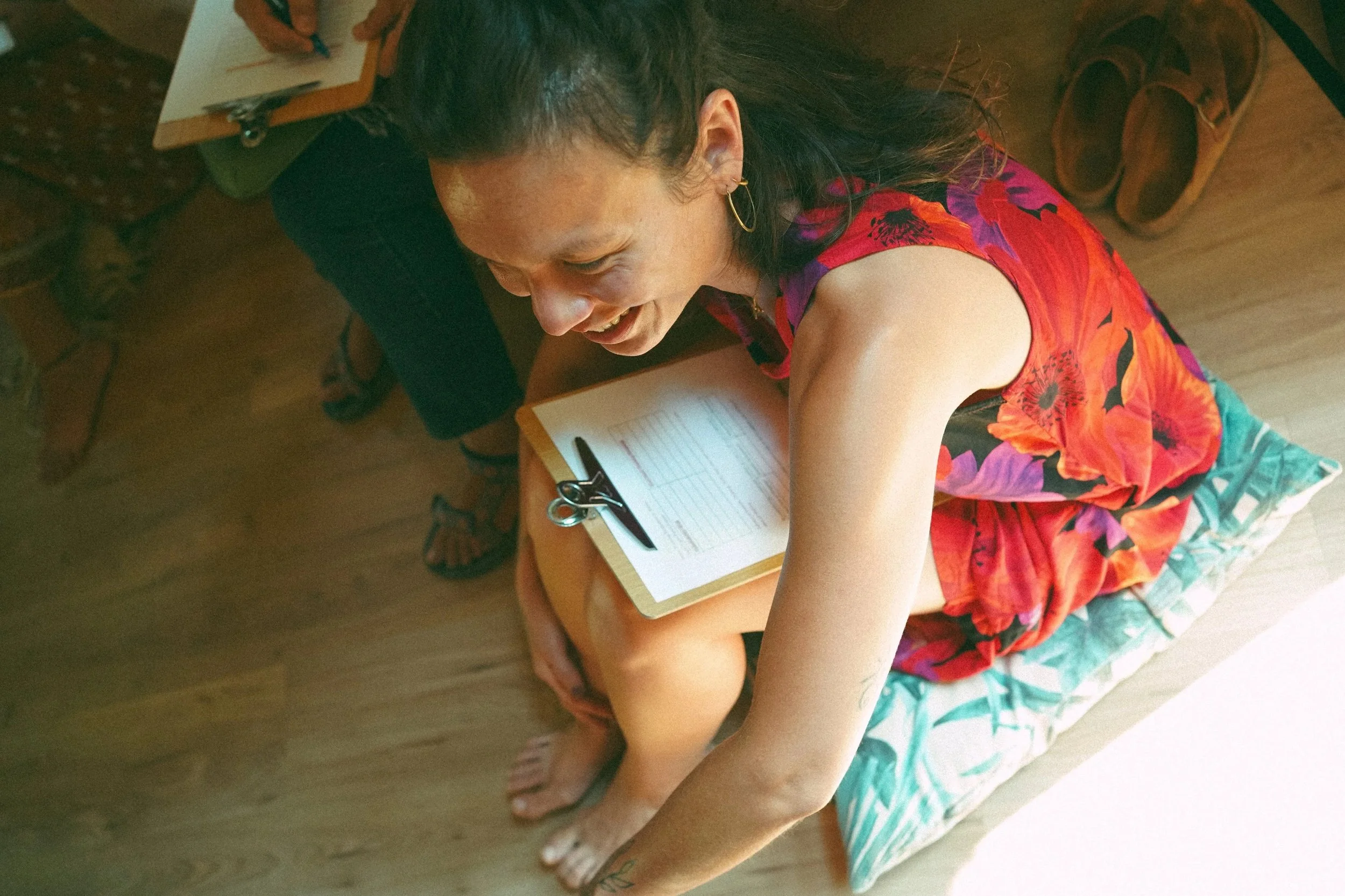 A woman sitting on a cushion on the floor, wearing a colorful floral dress, holding a clipboard with papers and a pen, smiling, and looking down. She has dark hair and is barefoot. There are shoes and a wooden floor visible in the background.