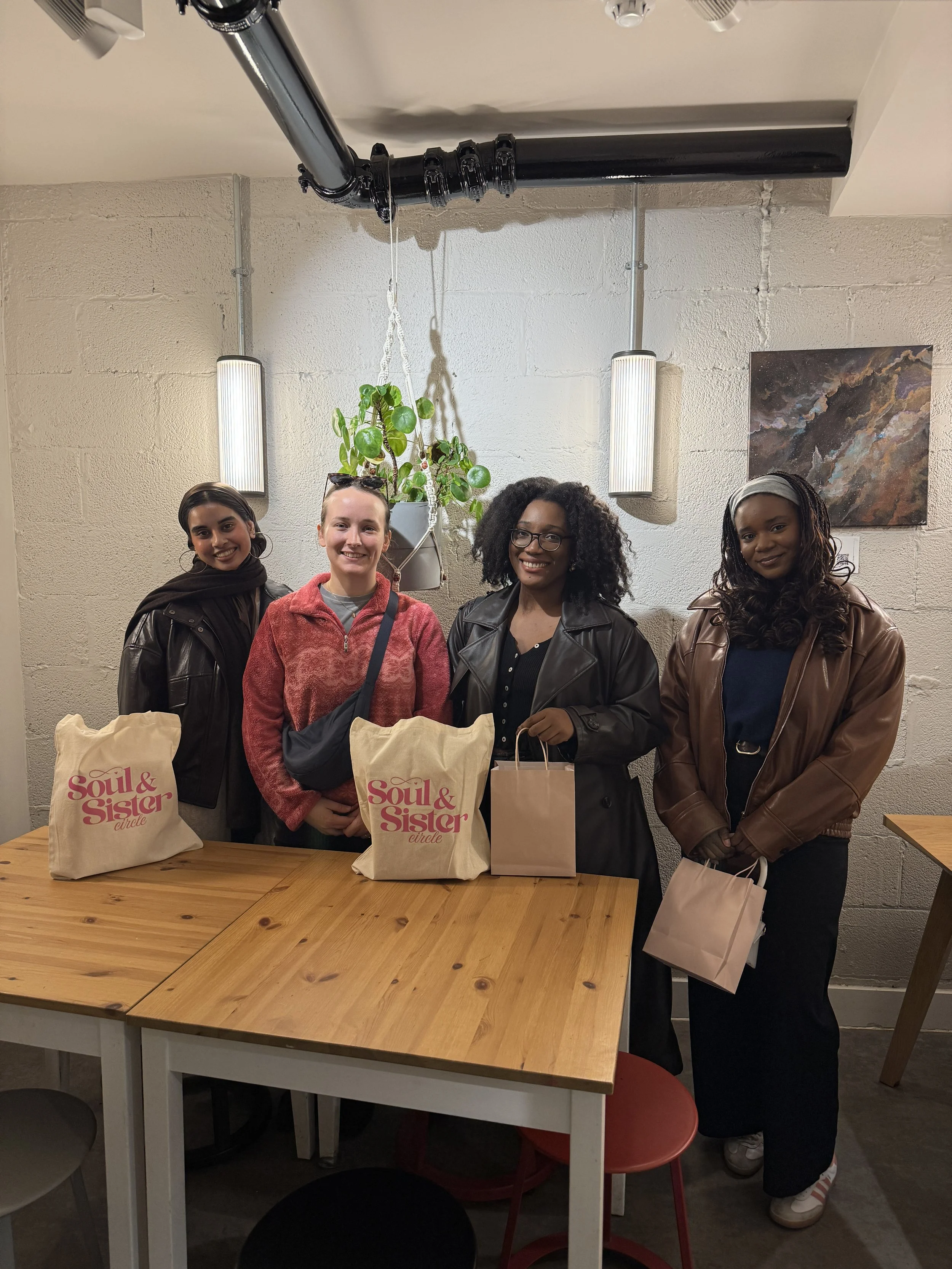 Four women standing behind a wooden table with gift bags, in a room with cream brick walls, modern light fixtures, and a hanging plant.
