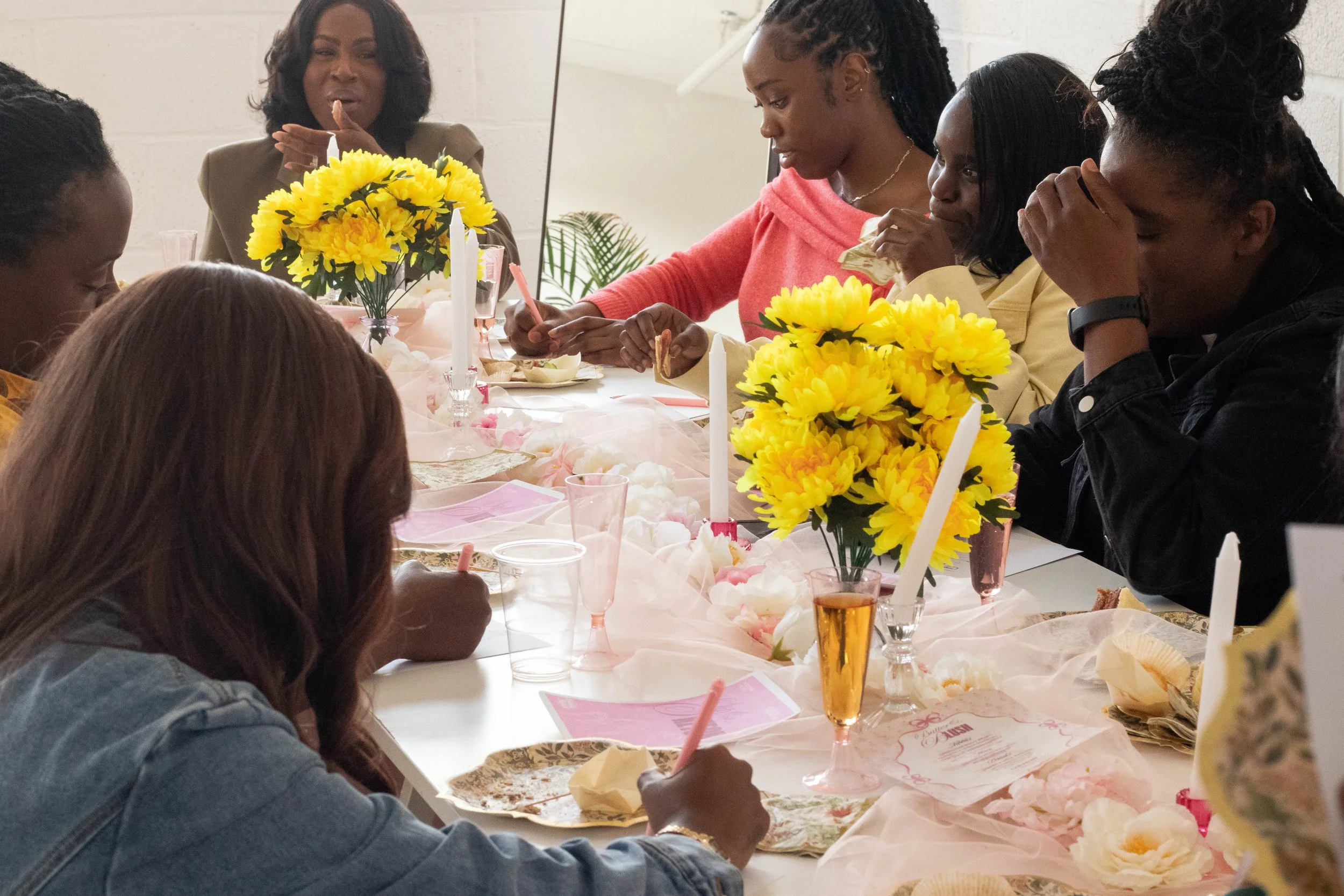 Women gathered around a table celebrating a special occasion, decorated with yellow flowers, pink napkins, and pink champagne glasses.
