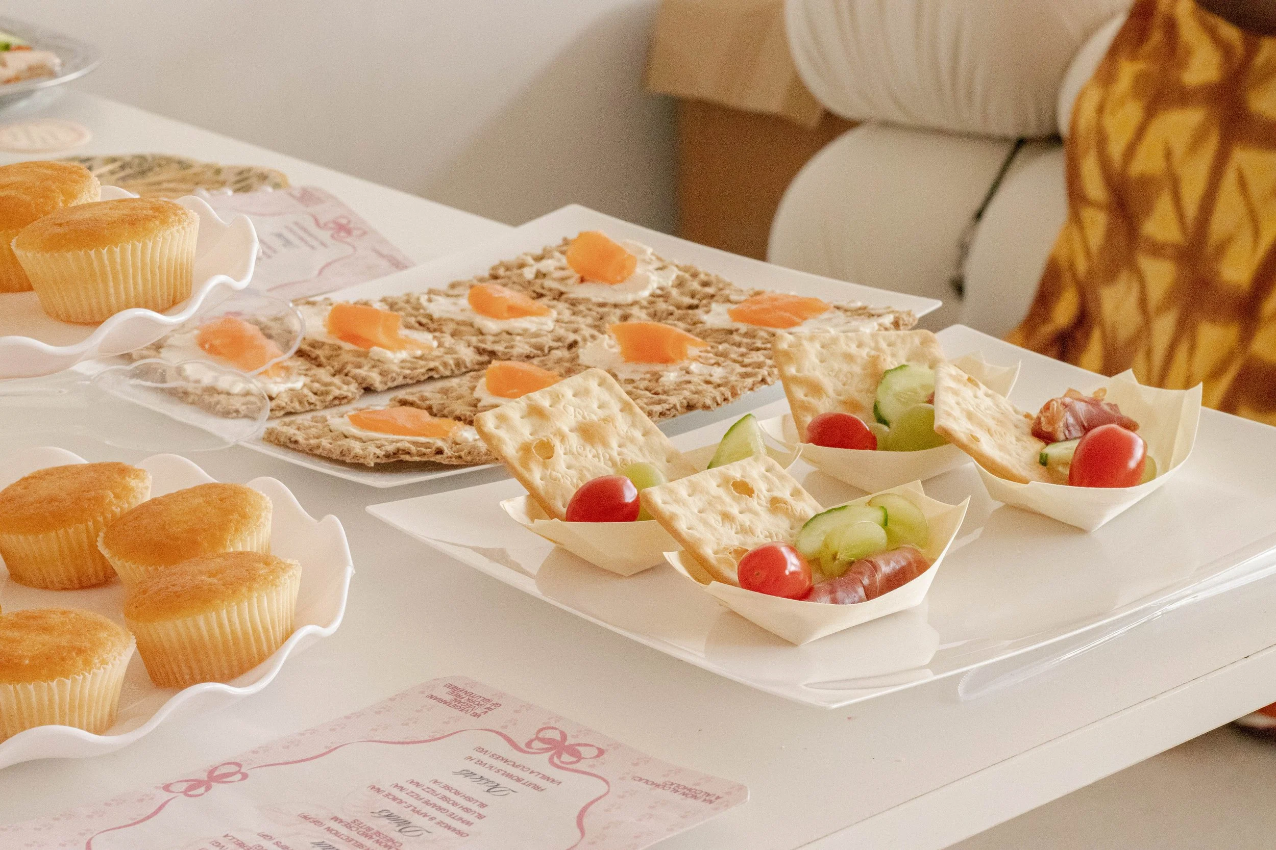 Assorted finger foods including mini muffins, crackers with cream cheese, cherry tomatoes, cucumber slices, and ham, arranged on white plates and bowls on a table.