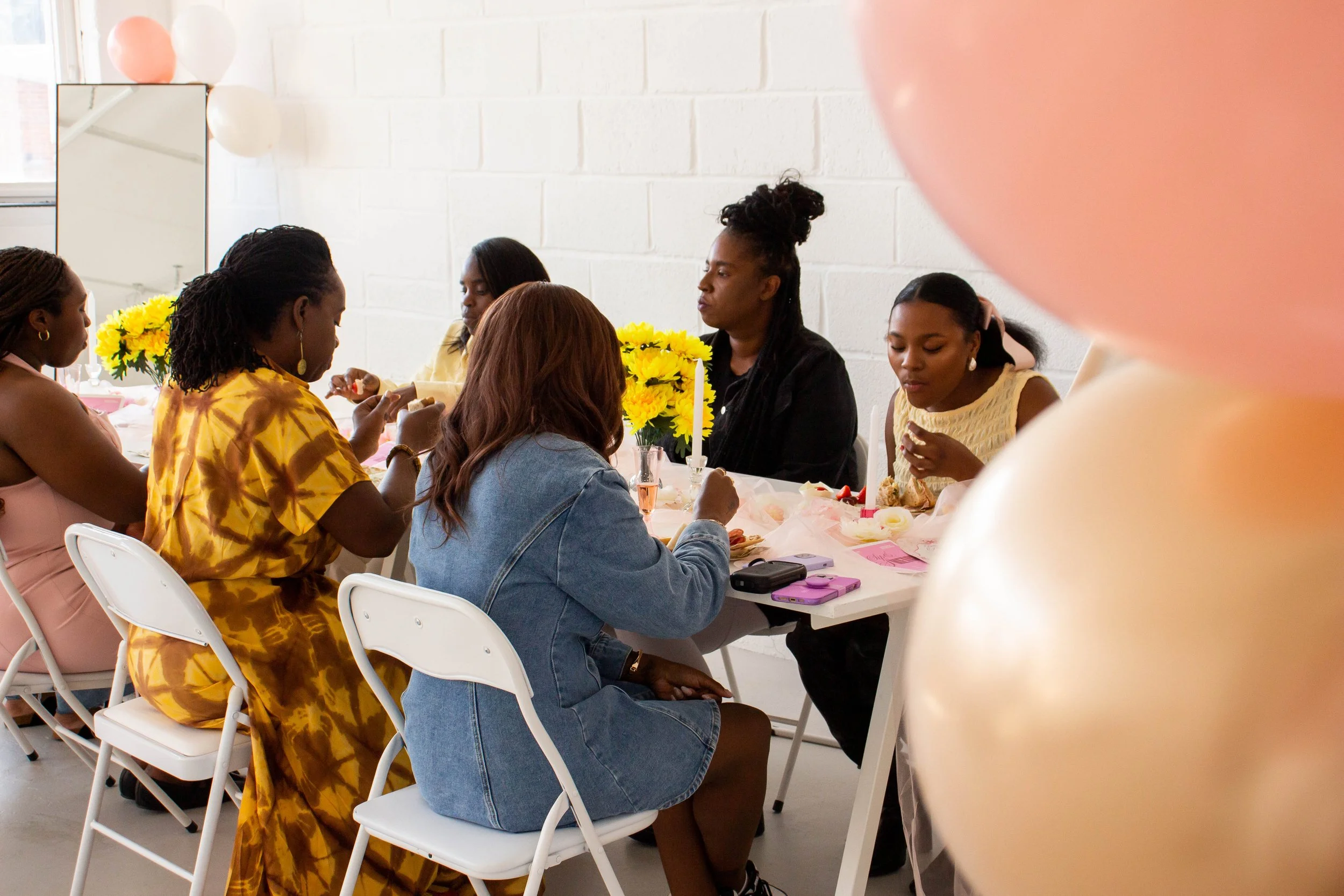 Group of women sitting at a decorated table having a celebration or gathering with pink, yellow, and peach balloons and flowers.