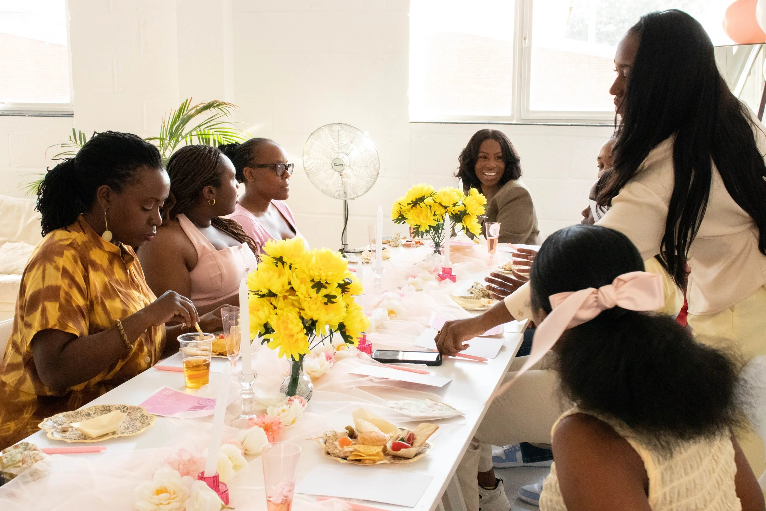 Women gathered around a decorated table with yellow flowers, celebrating indoors with food and drinks.