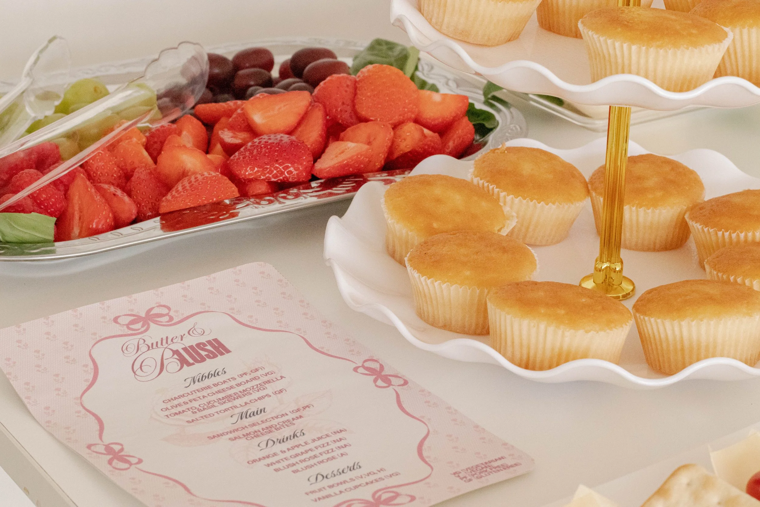 Assorted fresh fruit including strawberries, grapes, and melons on a silver platter, alongside a tray of cupcakes on a white tiered stand, and a pink menu card for a brunch event.