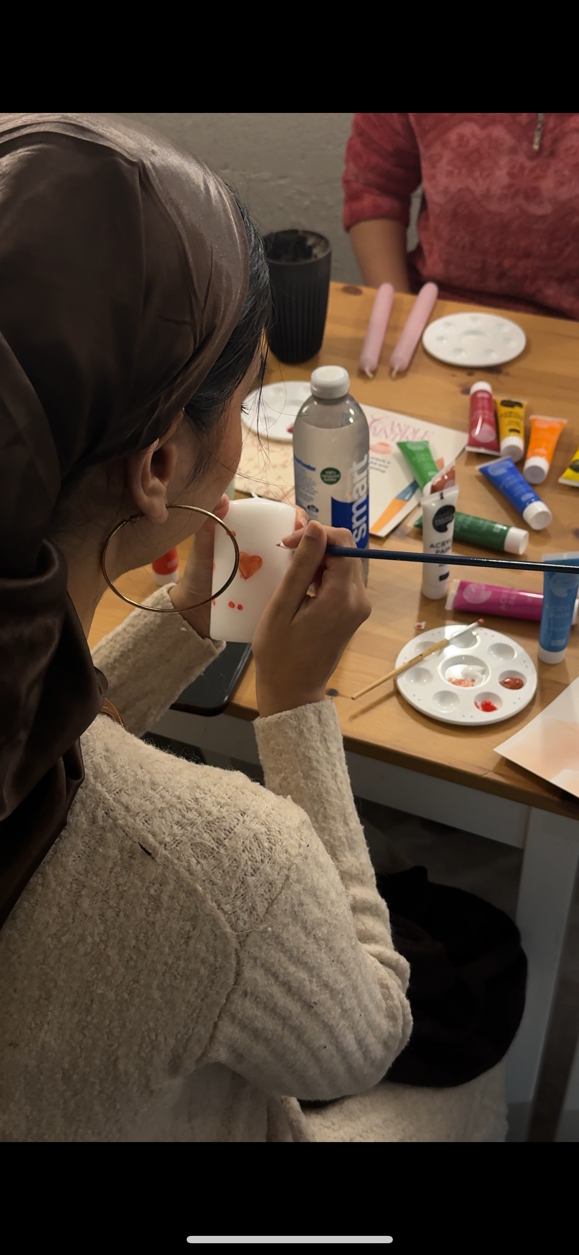 A woman painting a small white ceramic mug with red and orange designs at a table with art supplies, including paints, brushes, and palettes.