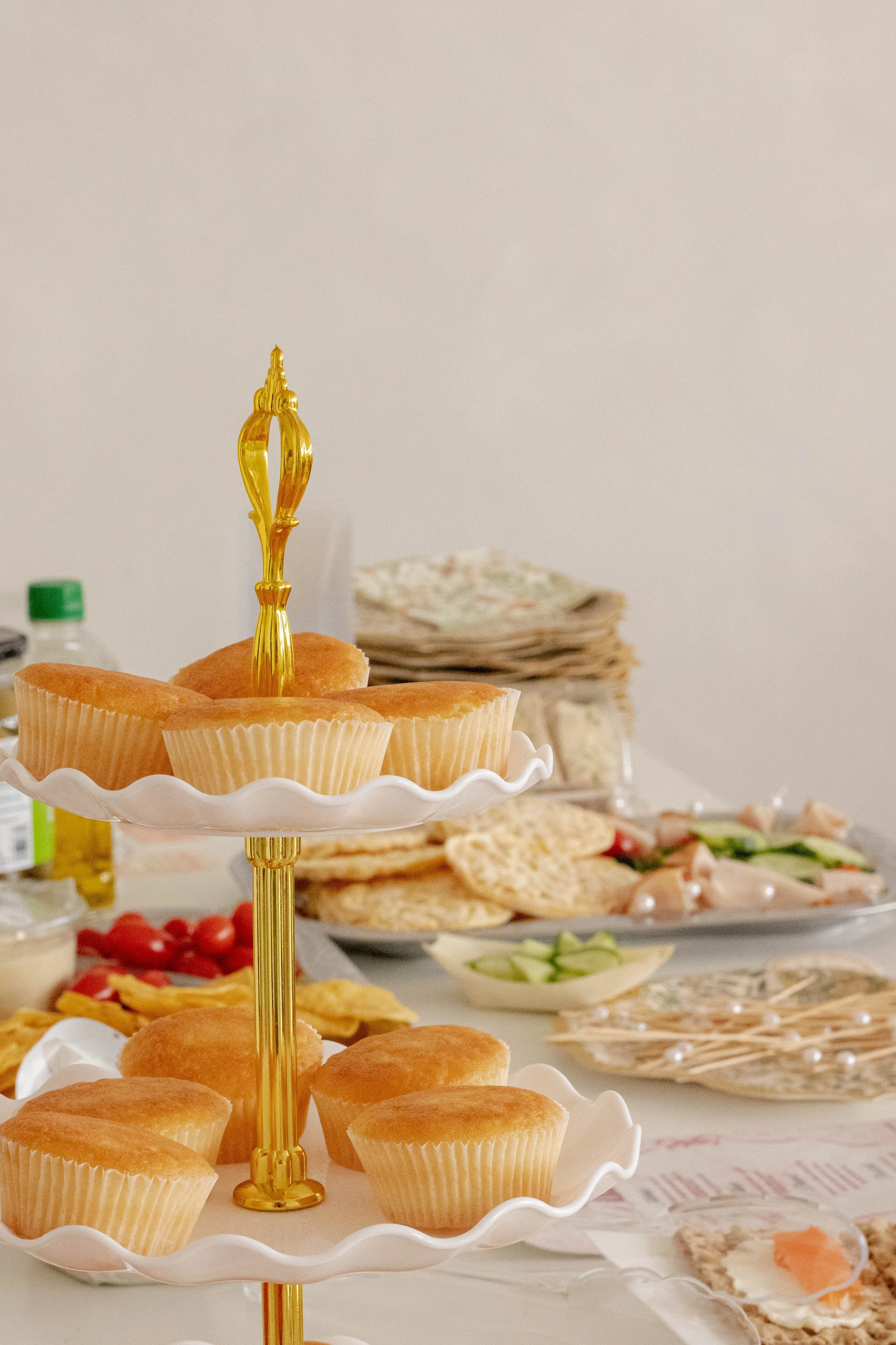 Plate of cupcakes on a two-tiered stand at a dessert table with various snacks, fruits, and napkins in the background.