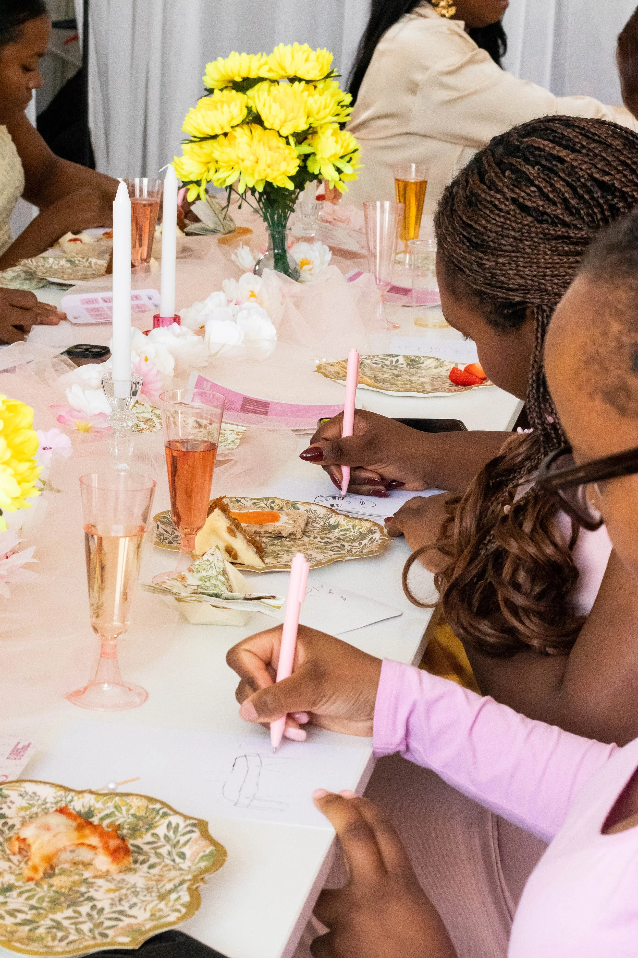 Women sitting at a decorated table, drawing on paper with pink pens, with floral and pink table decor including a centerpiece of yellow flowers, surrounded by plates with food and pink glasses.