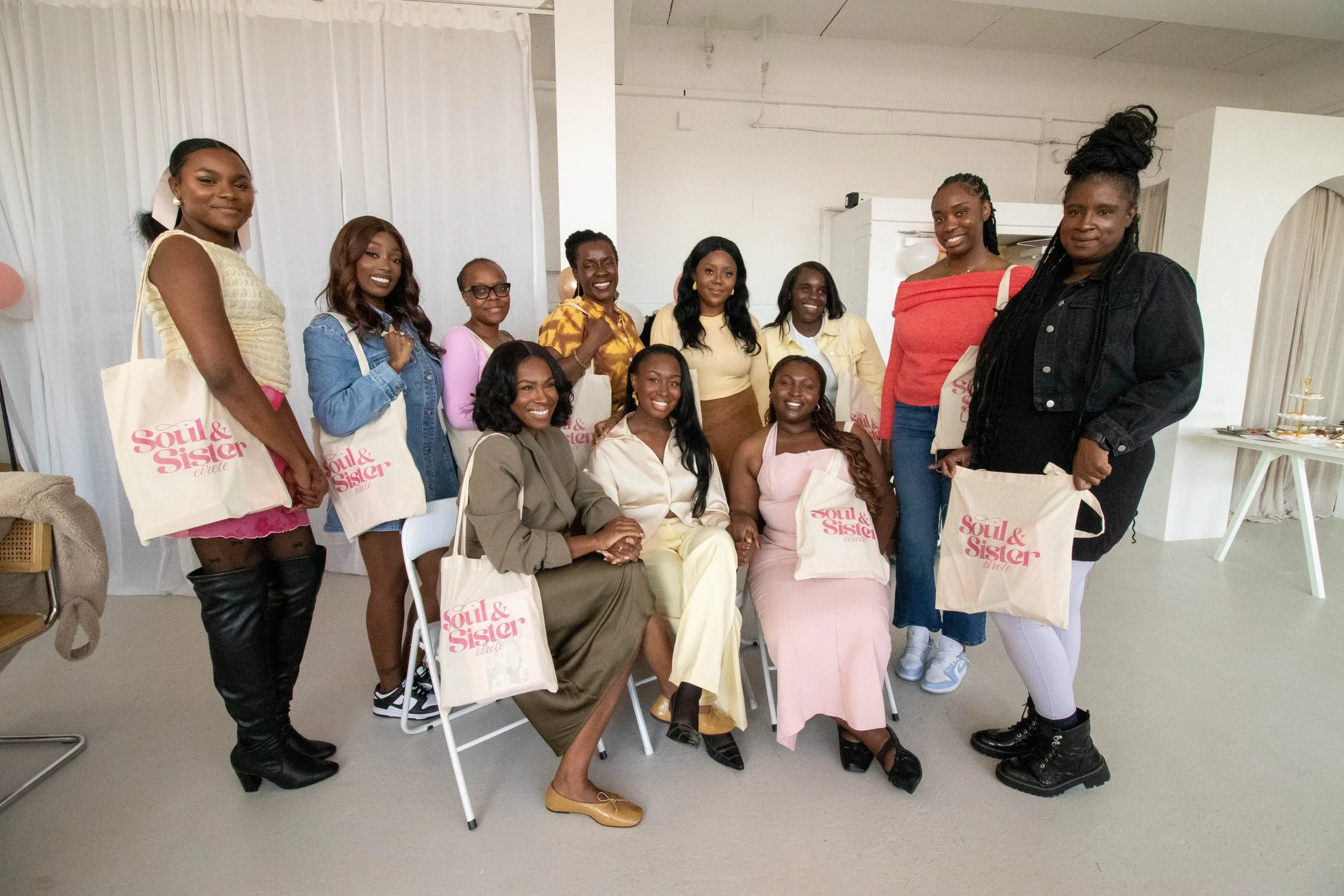 A group of women posing together indoors, many holding tote bags with the words 'Soul & Sister' printed on them, smiling, dressed in casual and semi-formal attire.