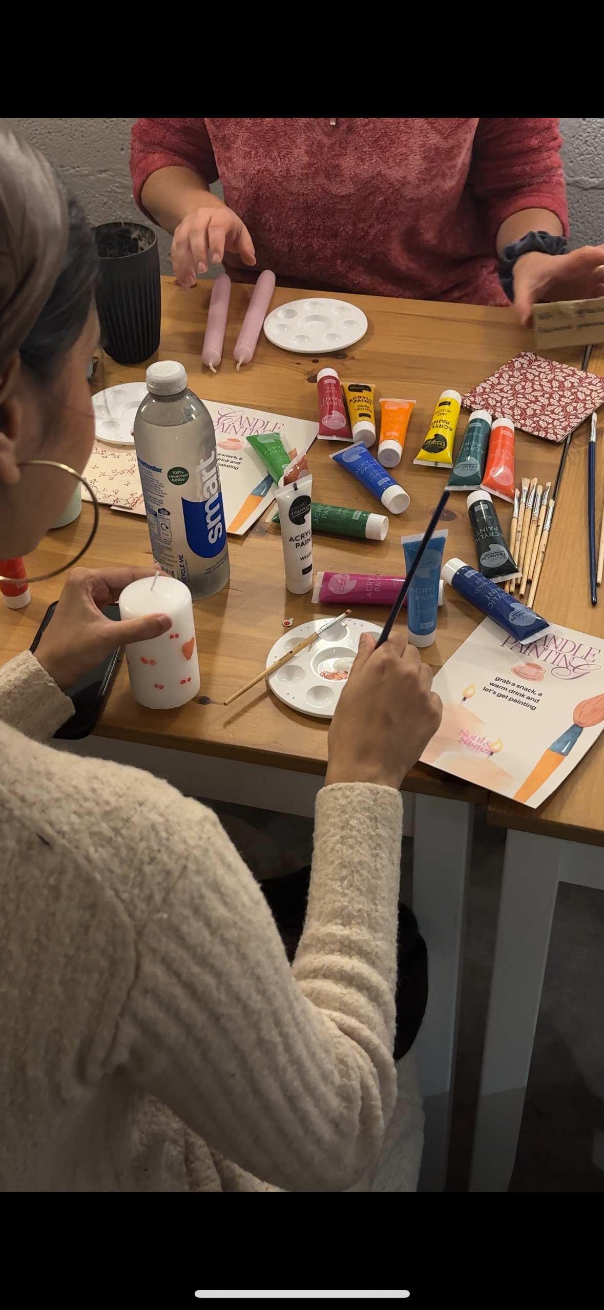 People at a table decorating candles with paint and brushes, surrounded by supplies such as paint tubes, brushes, and candle-making kits.