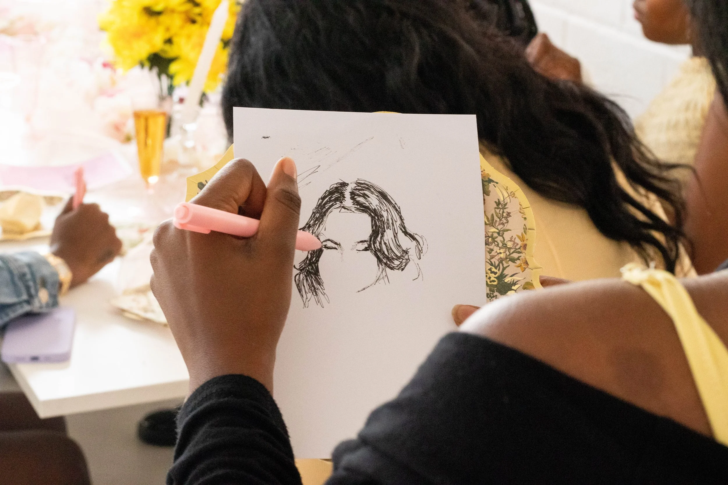 Person drawing a sketch of a woman's face on a white paper at a gathering with other people, flowers, and plates on the table.