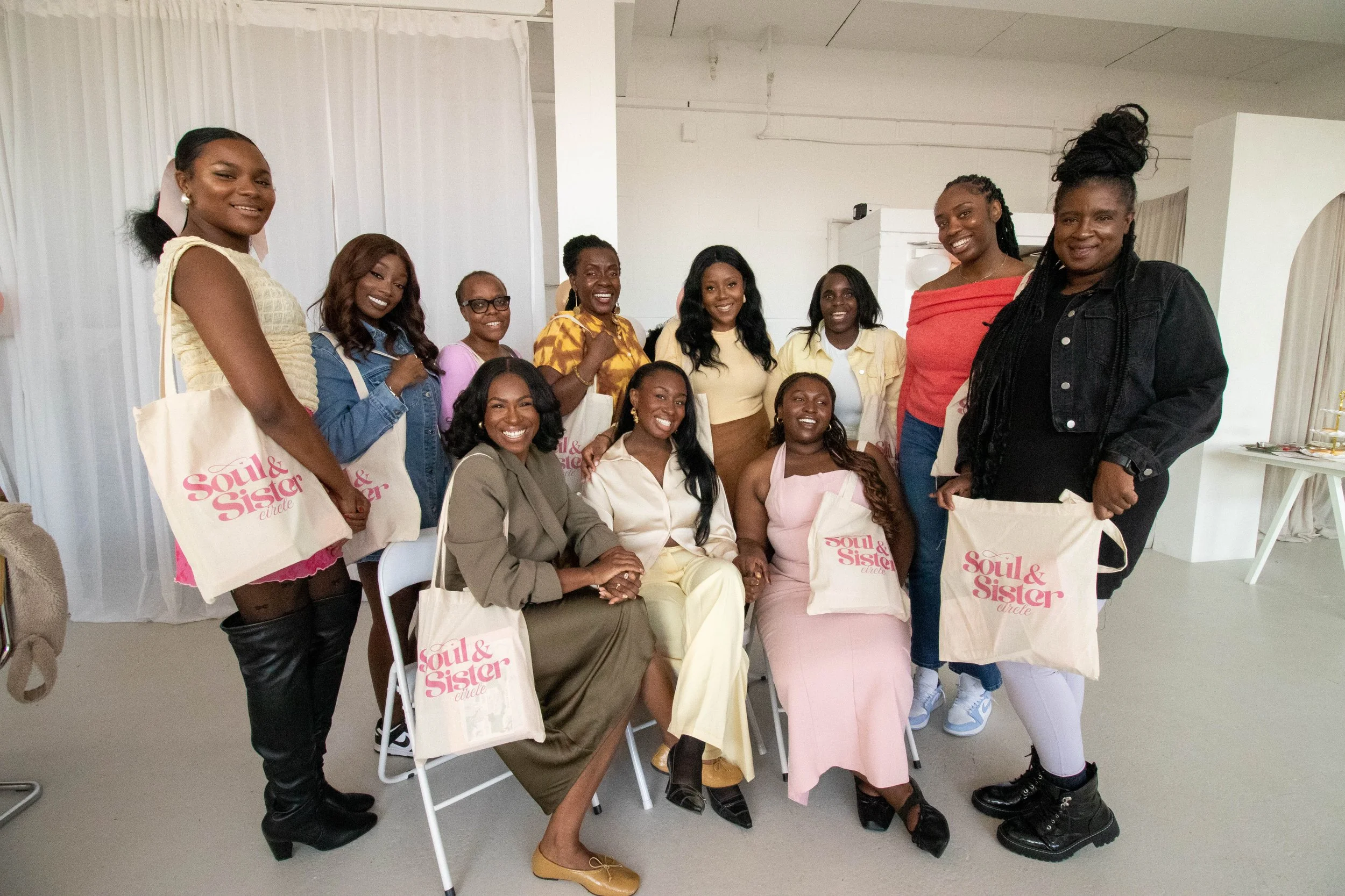 Group of women at a gathering, holding tote bags that say 'Soul & Sister Circle,' indoors with a white background and curtains.