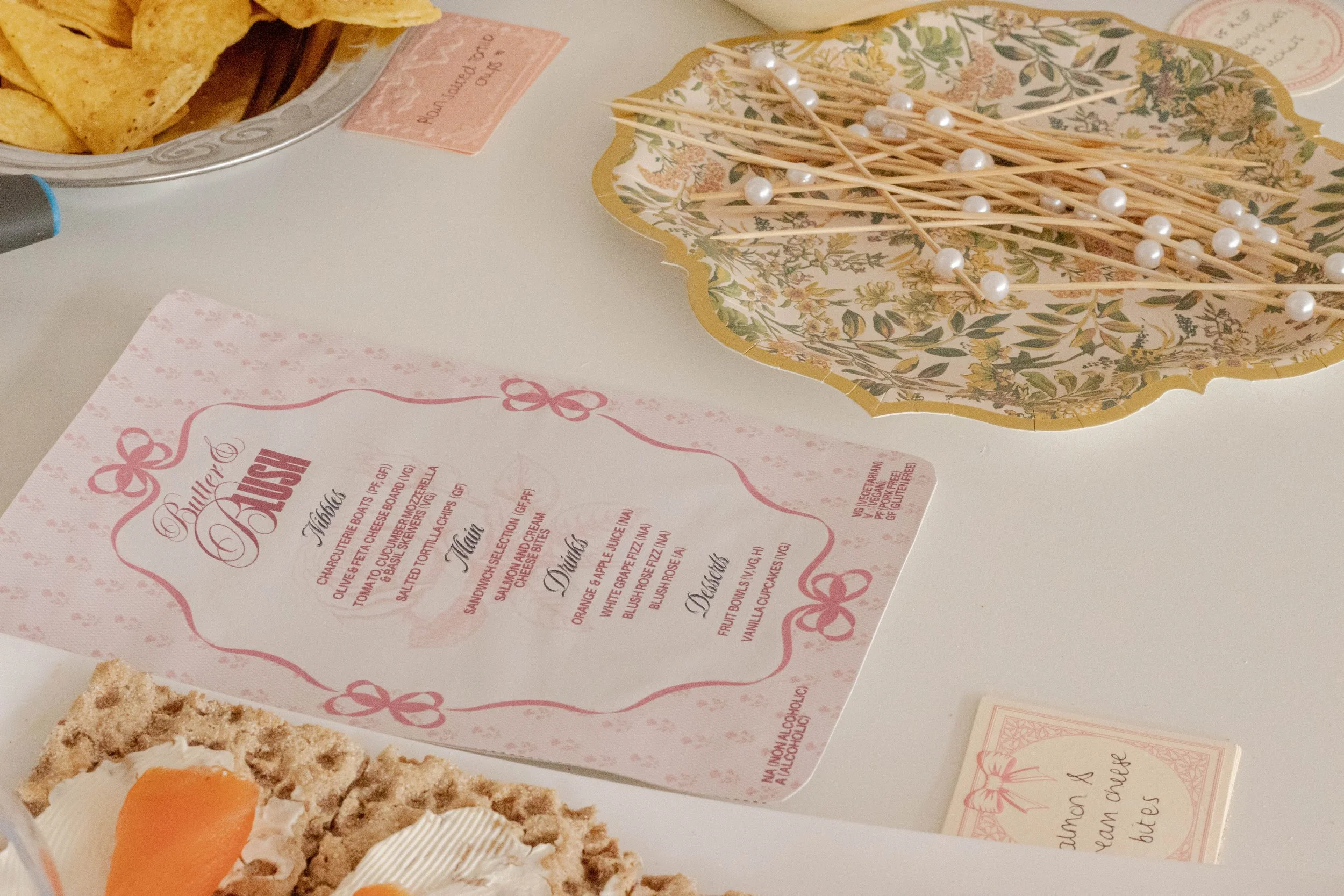 A table with a menu titled 'Balletto & Burrow' listing drink, main, and dessert options. A floral tray with pearl-topped toothpicks, a partially visible slice of cake with orange and cream toppings, and a bowl of tortilla chips are also on the table.