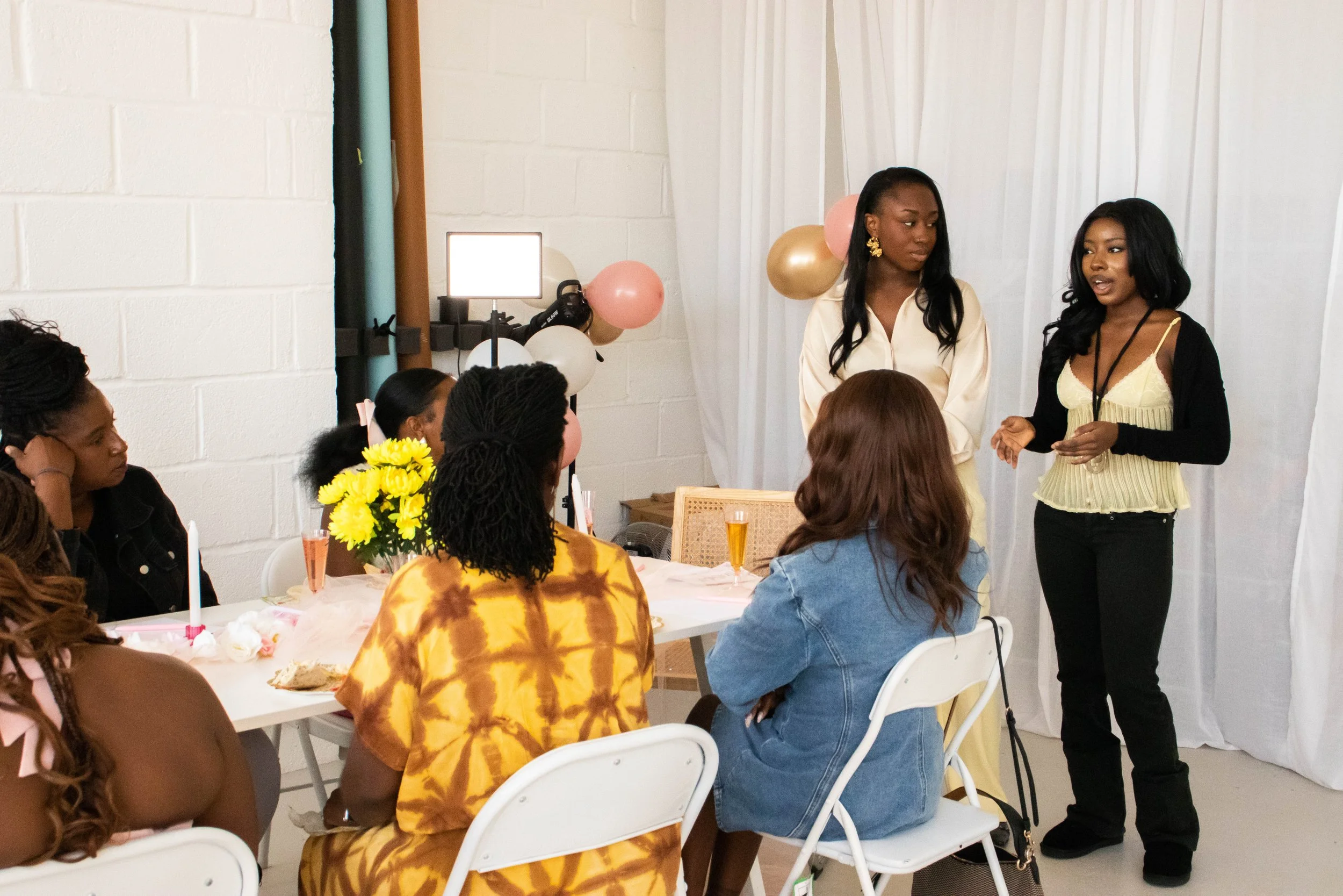 A group of women sitting around a table at a social gathering, with two women standing and speaking to the group. The table has yellow flowers and candles, with balloons in the background.