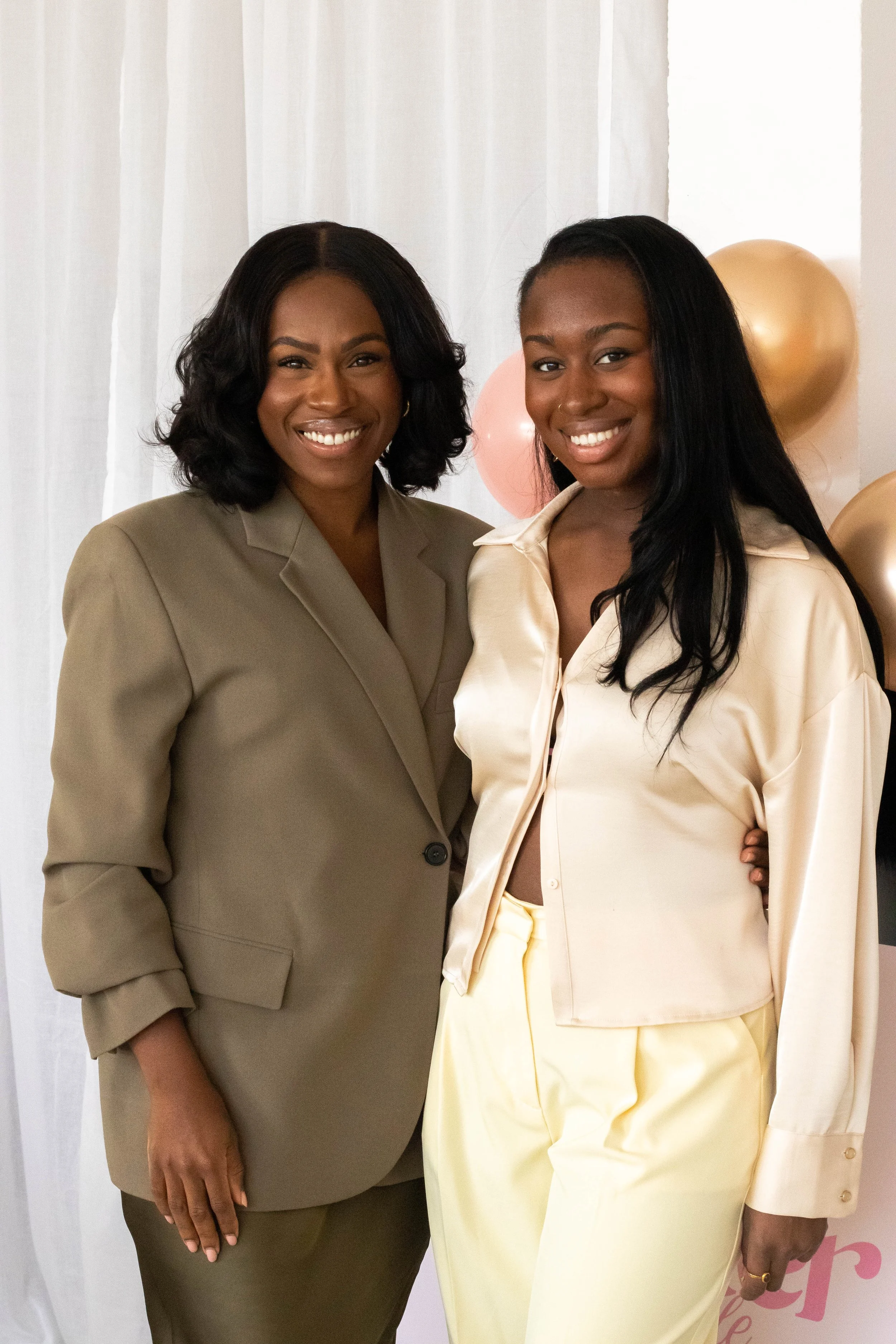 Two women standing together indoors, smiling, with balloons in the background.